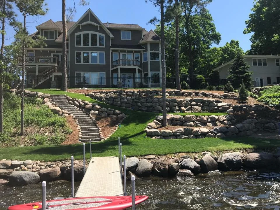 A large, multi-story house with a landscaped yard, stone retaining walls, and a dock leading to a body of water in the foreground, under a clear blue sky.