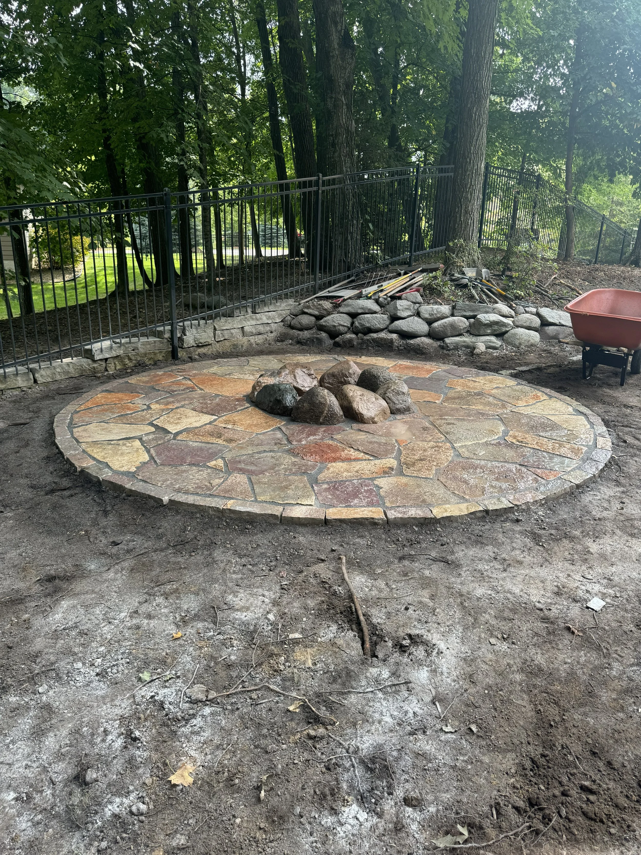 A circular stone patio with rocks in the center, surrounded by dirt, with a black fence, trees, and garden tools in the background.