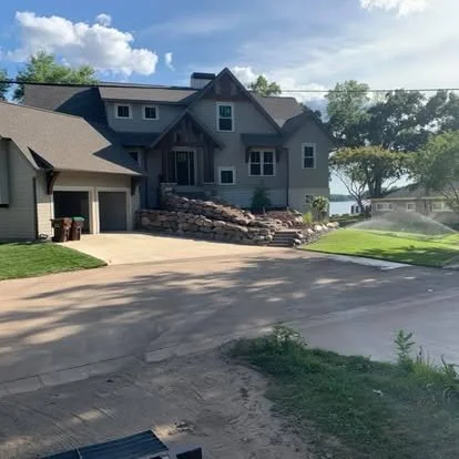 Residential house with a stone staircase leading to the front door, surrounded by a driveway and green lawn, under a partly cloudy sky.