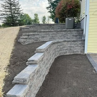 A tiered stone retaining wall next to a house with an air conditioning unit on top, surrounded by dirt and some grass, with trees in the background.