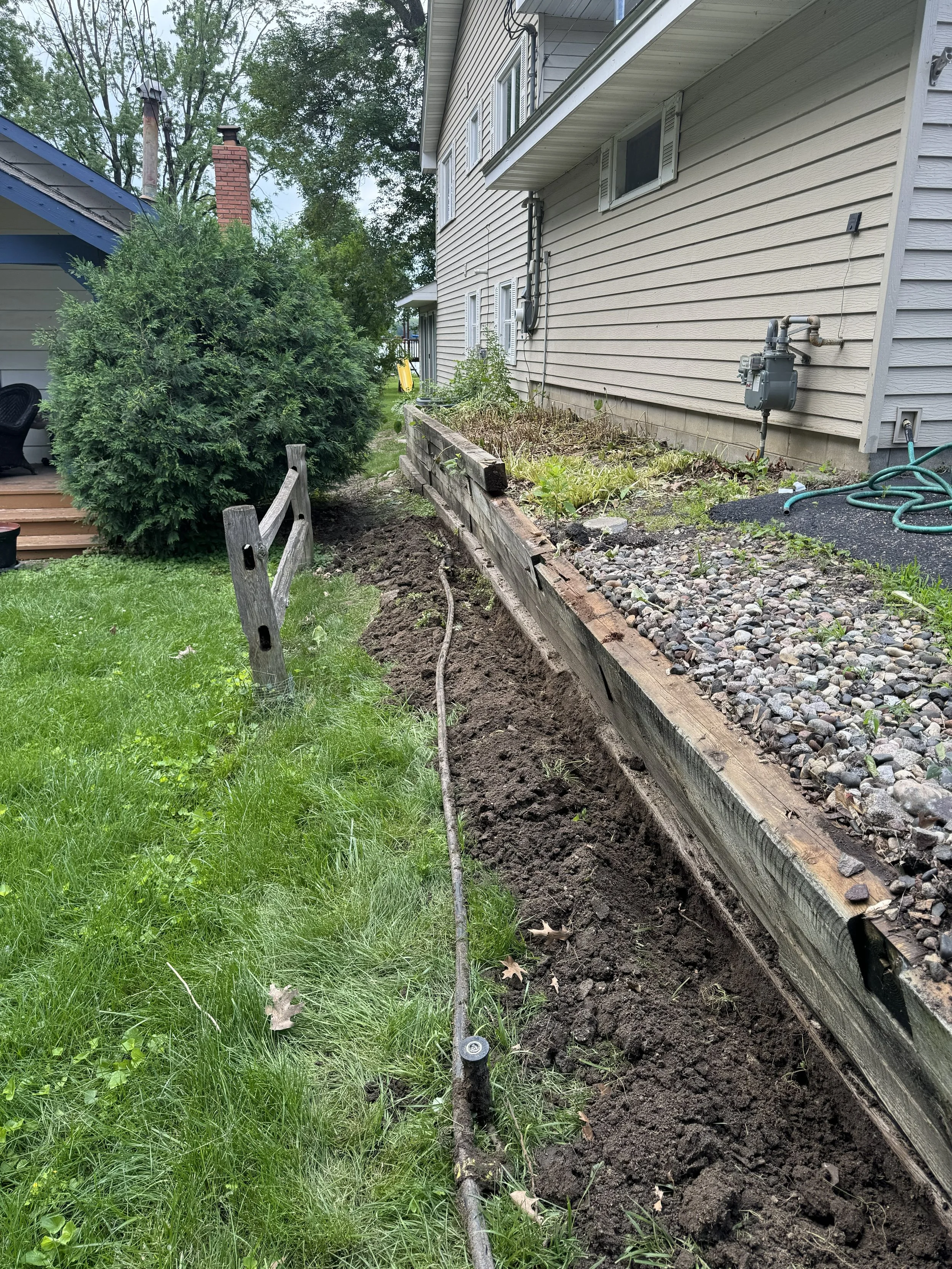 Side yard of a house with a fresh trench dug along the foundation, garden bed with rocks, green lawn, large bush, and hoses.
