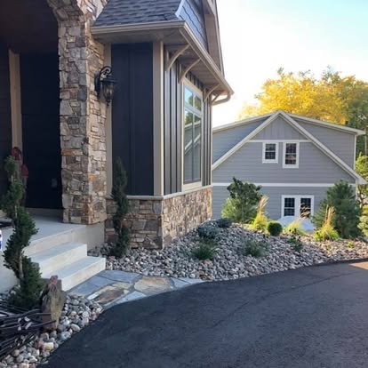 A side view of a house's front entrance with stone steps, dark blue siding, large window, and landscaped front yard with rocks and small plants.
