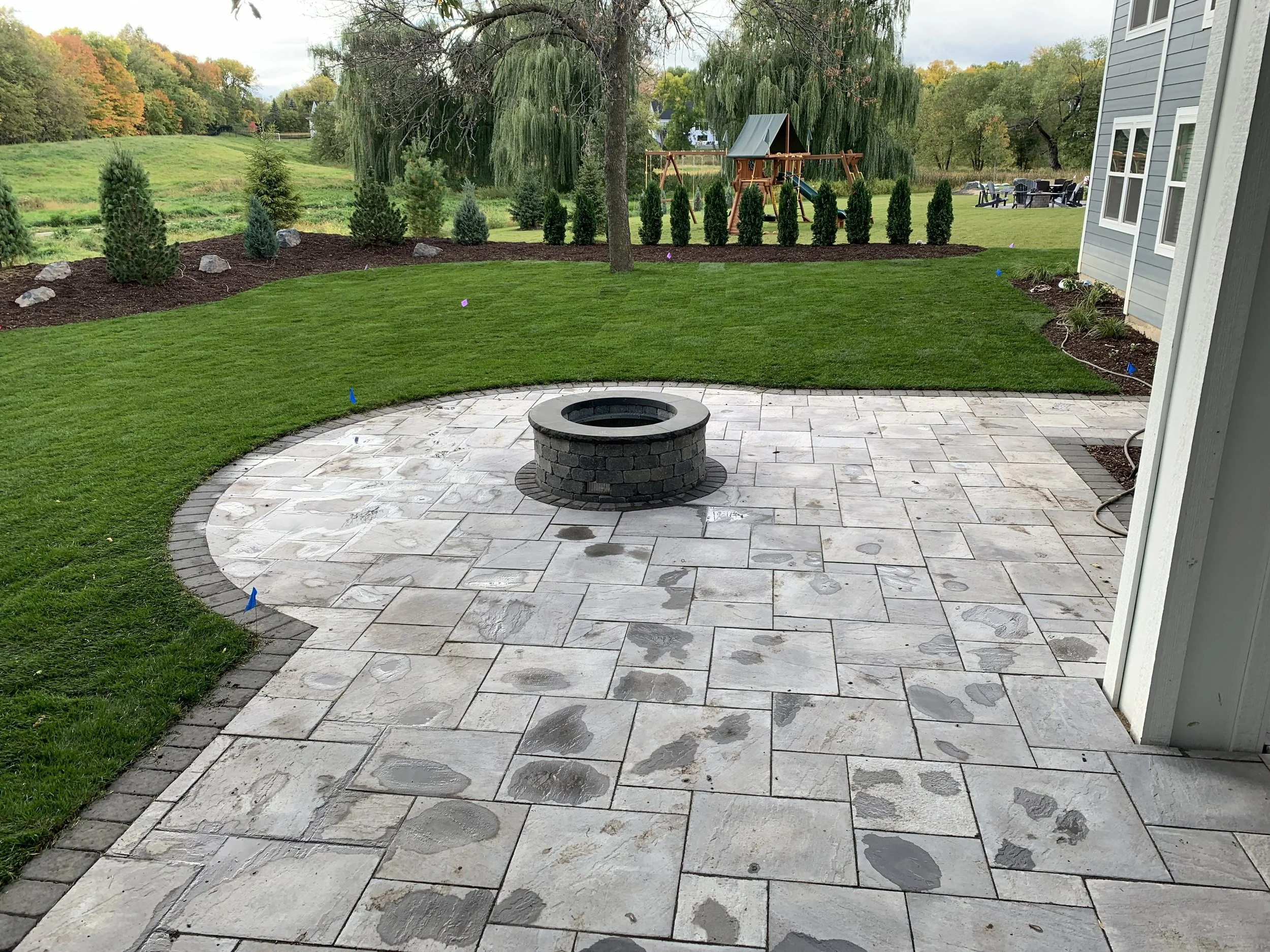 Patio with stone pavers, a circular fire pit in the center, surrounded by grass and landscaped yard with trees and a children's playset in the background.