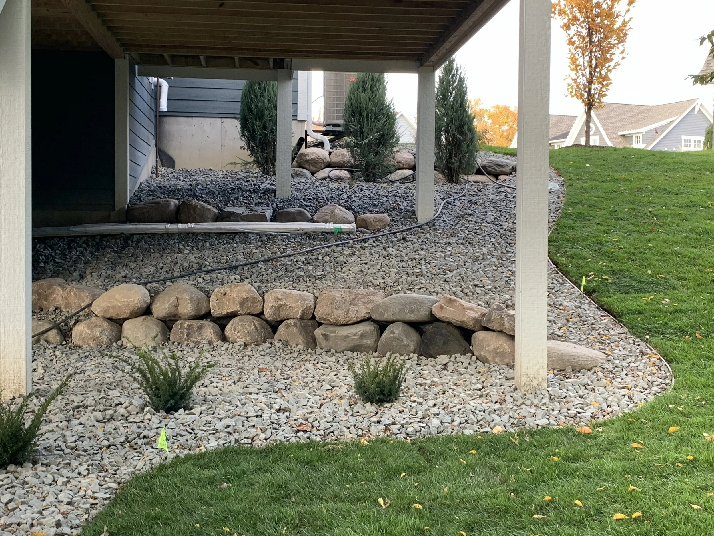 A landscaped area underneath a porch with a bed of small rocks, larger rocks, small bushes, and a cable running across the rocks.