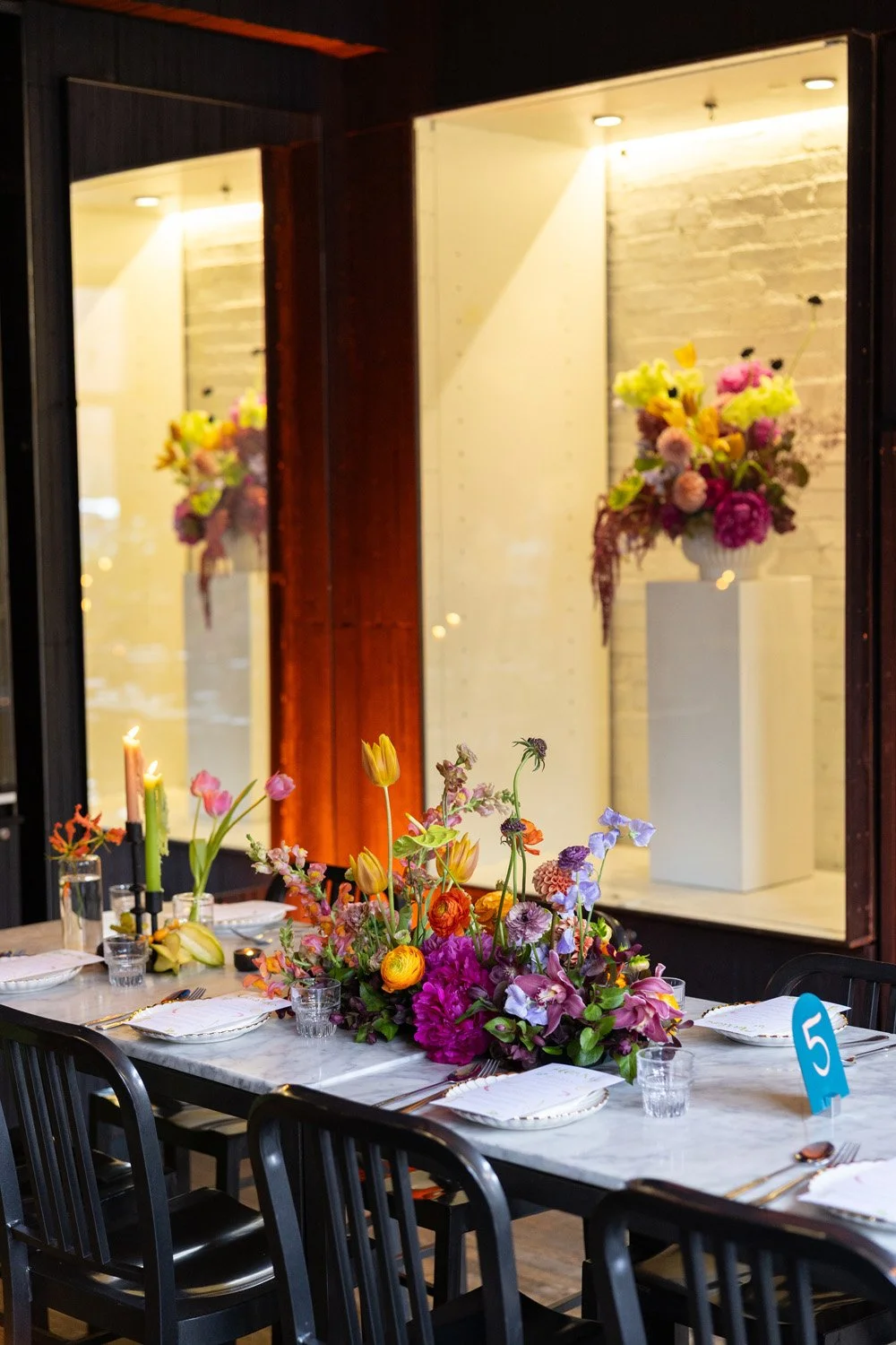 A banquet table decorated with an elaborate centerpiece of colorful flowers, candles, and dining ware, with a large floral arrangement in a display case in the background.