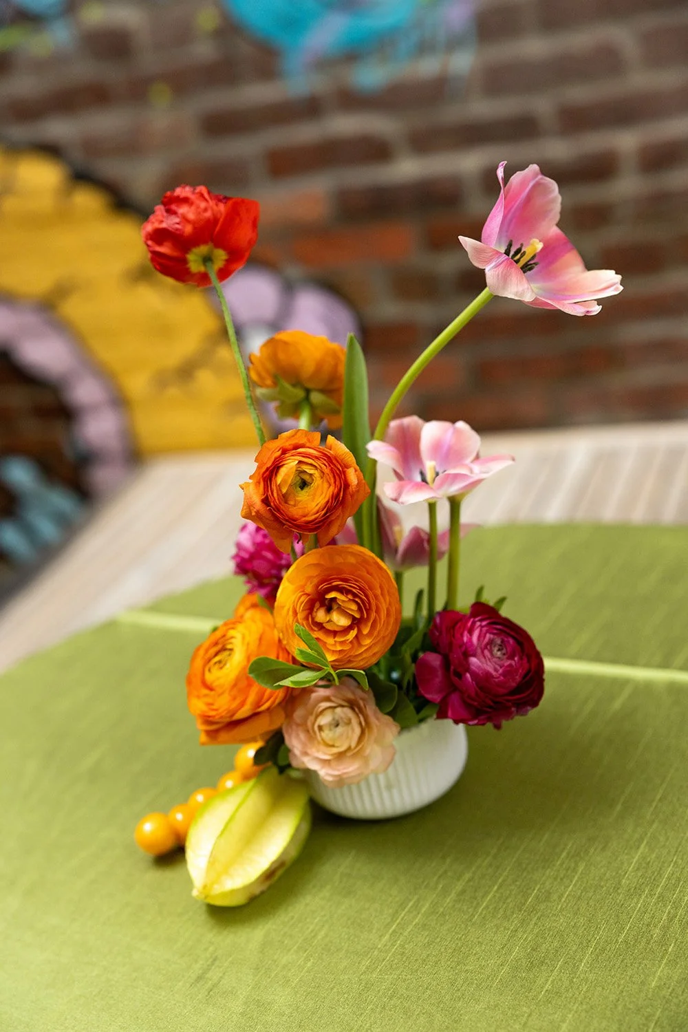 Colorful flower arrangement in a white vase with pink, orange, yellow, and red flowers on a green tablecloth, against a brick wall background.
