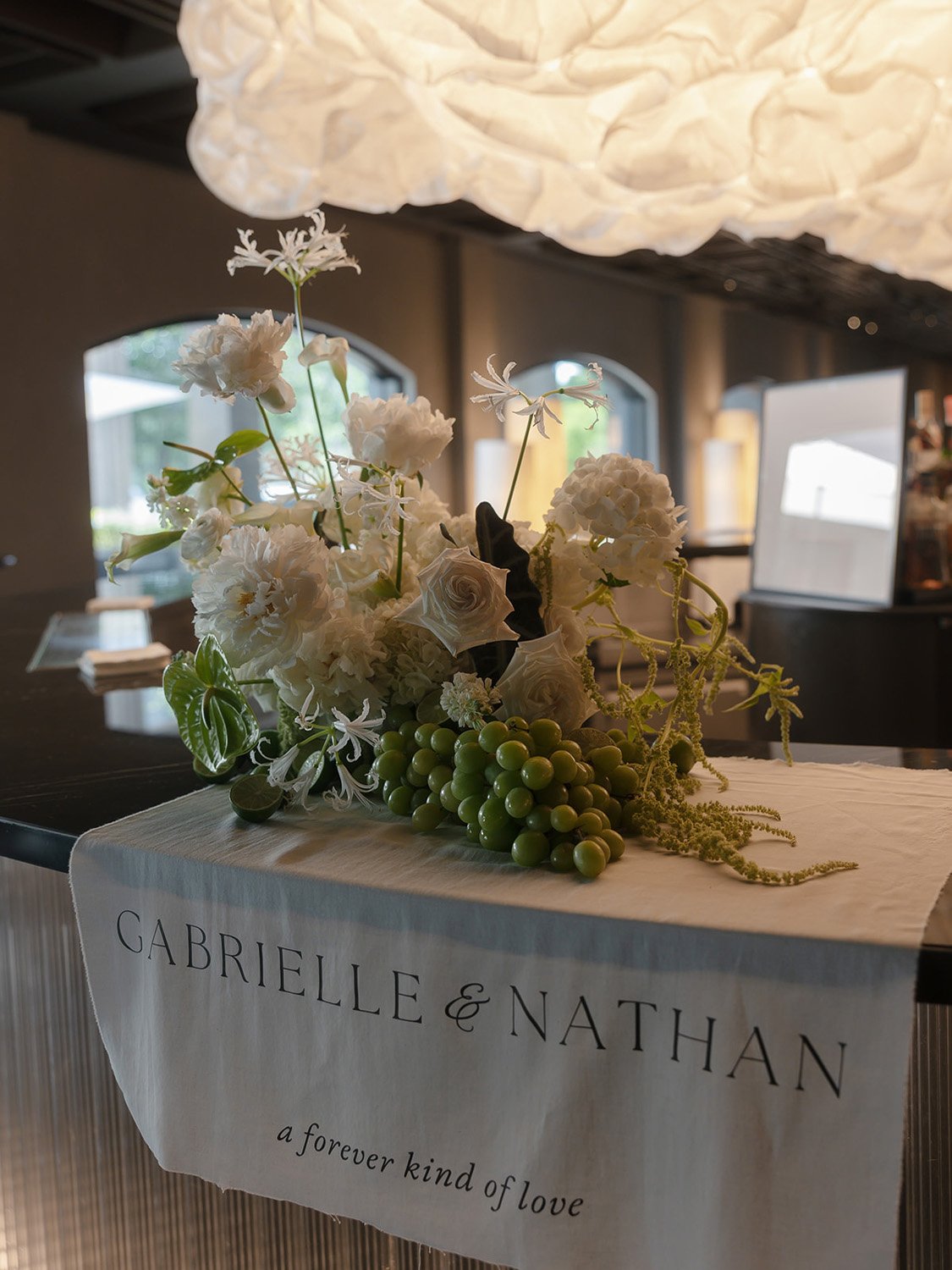 A floral arrangement with white flowers and green grapes, placed on a table with a cloth that has the names Gabrielle and Nathan and a message about love. The background shows a room with windows and a decorative ceiling light.