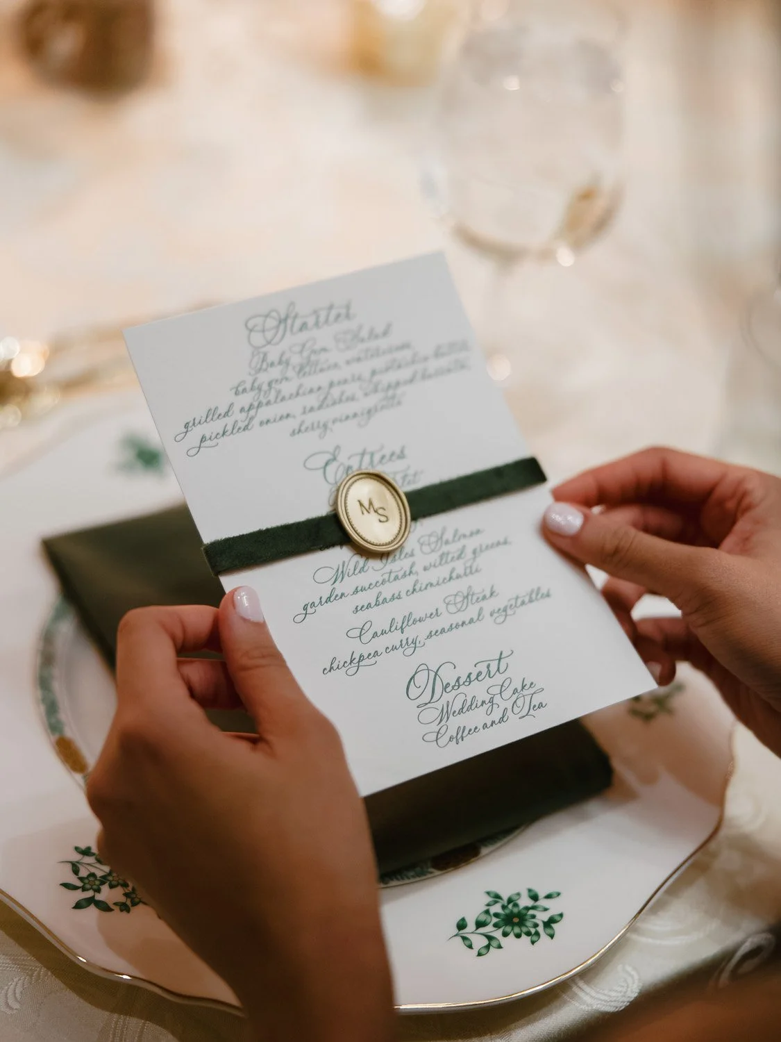Person holding a wedding menu card with a black ribbon and a gold crest marked with initials, on a decorated table setting with glasses and a floral plate.