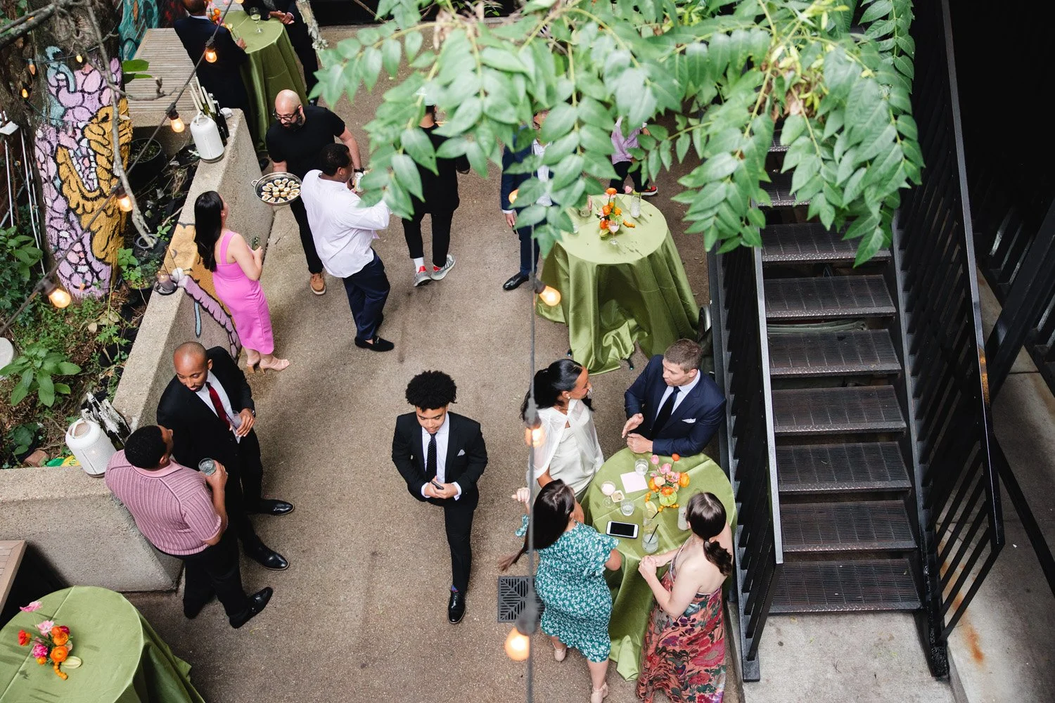 People socializing at a party in an outdoor venue, with some standing near tables with green tablecloths and floral centerpieces, and others engaged in conversation. There are string lights hanging above.