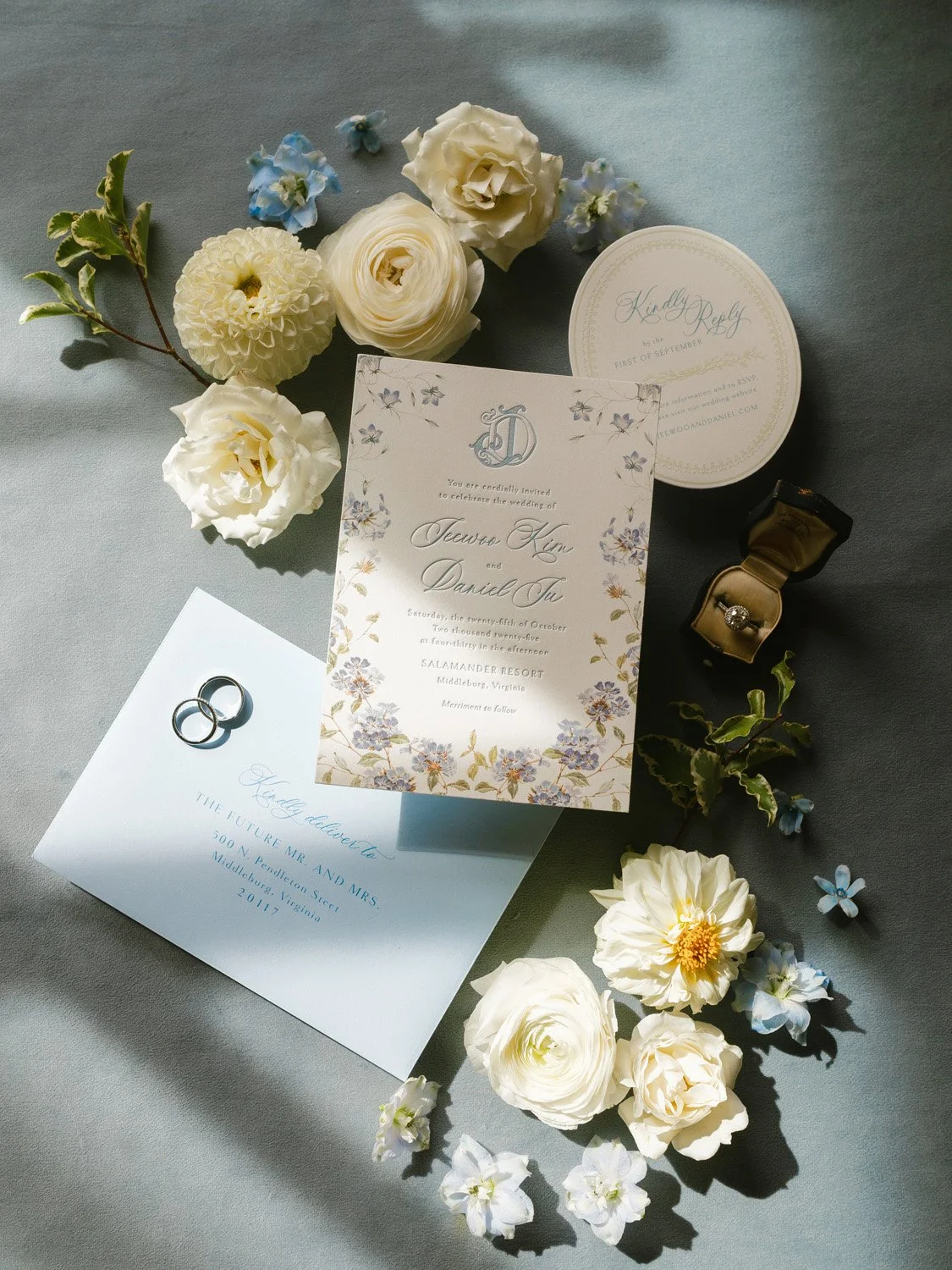 Wedding invitation, engagement rings, wedding band, and flowers on a gray surface.
