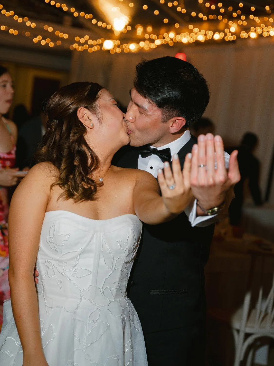 A bride and groom kiss during their wedding reception, with the bride in a white strapless dress and the groom in a tuxedo. The bride is holding the groom's hand, showing her wedding ring, and guests are visible in the background under string lights.