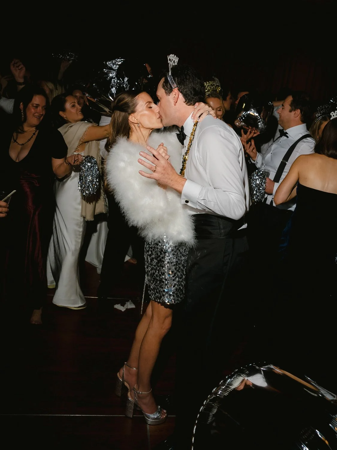 Couple kissing at a celebration party with others wearing festive accessories and holding balloons in the background
