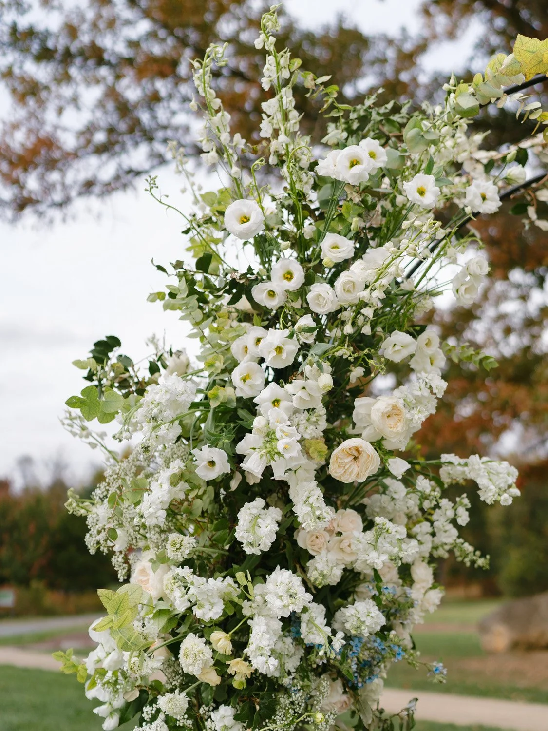 A vertical floral arrangement with white flowers and greenery, set outdoors with blurred trees and grass in the background.