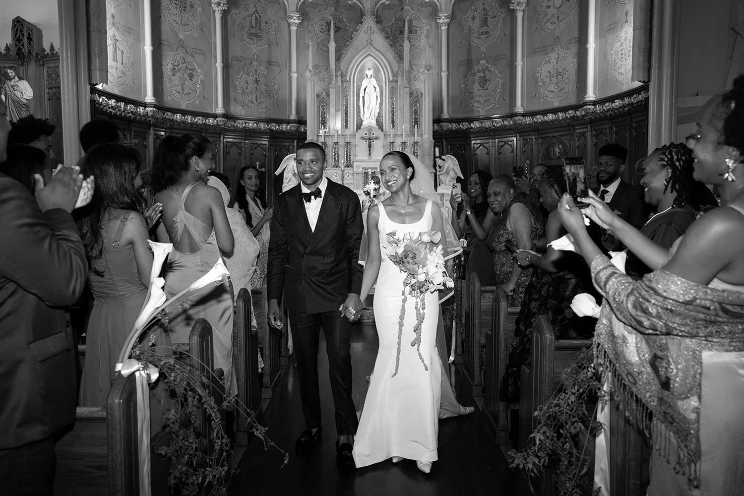 A couple in wedding attire walking down the aisle of a church, surrounded by happy guests. The bride is holding a bouquet and wearing a wedding dress, while the groom is in a tuxedo.