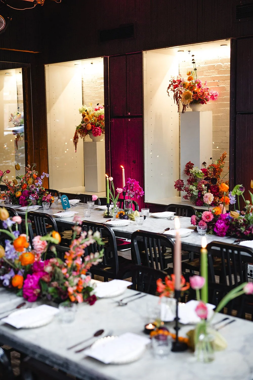 Formal dining setup with a long table decorated with colorful floral arrangements, candles, and place settings, with wall-mounted displays of flower arrangements