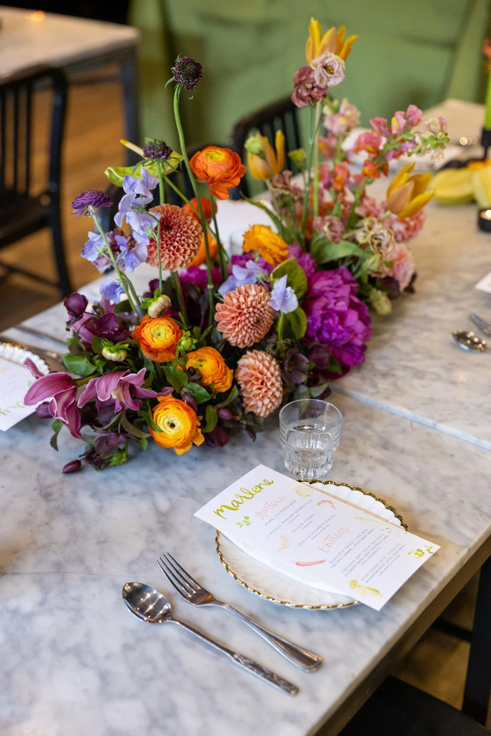 A dining table decorated with a large colorful floral centerpiece, a printed menu on a white plate, a small glass of water, a fork and a spoon, with part of a napkin and other tableware visible in the background.