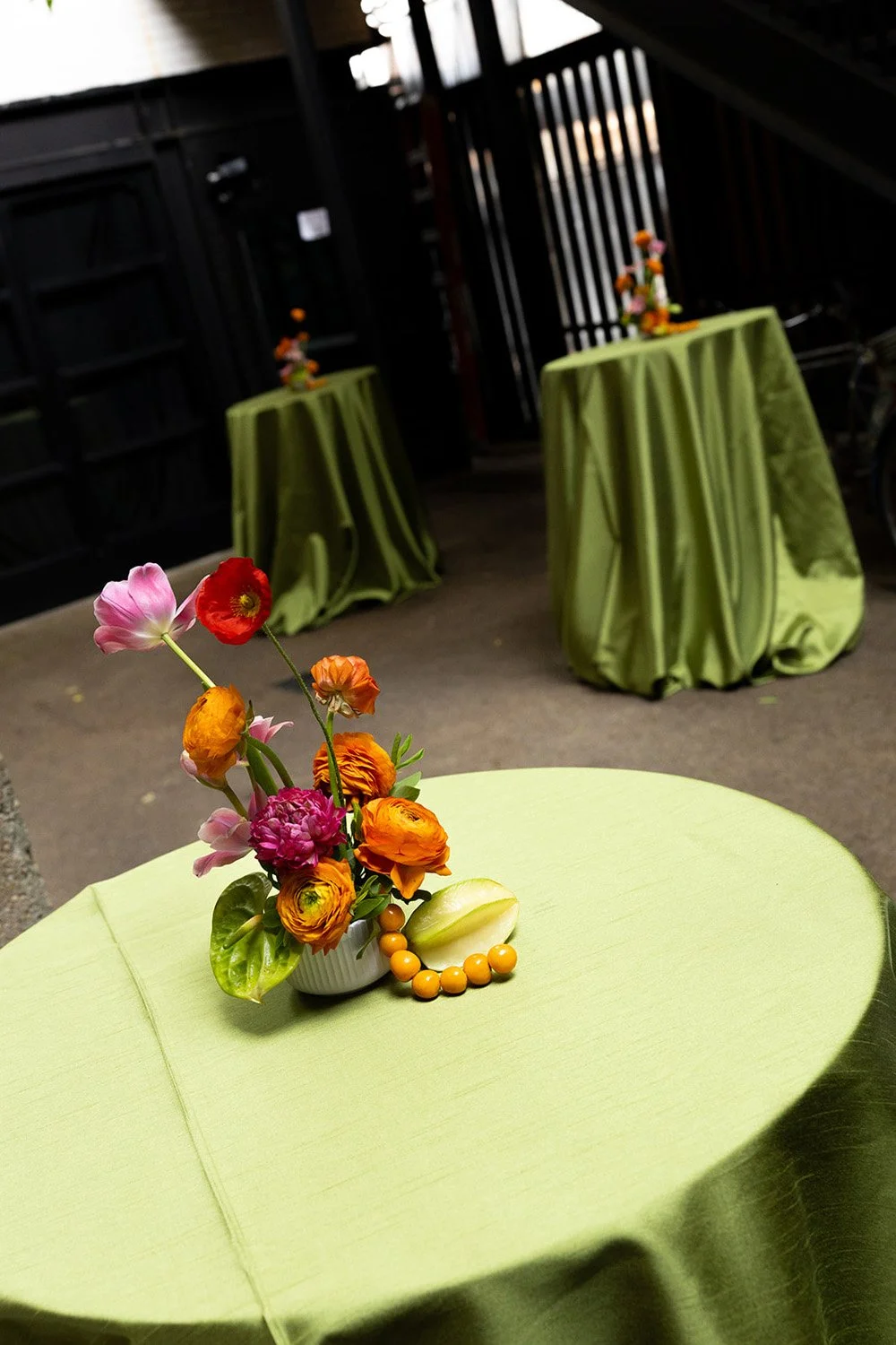 A floral centerpiece with pink, red, orange, and purple flowers in a white vase on a light green tablecloth. In the background, there are two more tables covered with matching green tablecloths, each with small floral arrangements. The setting appear
