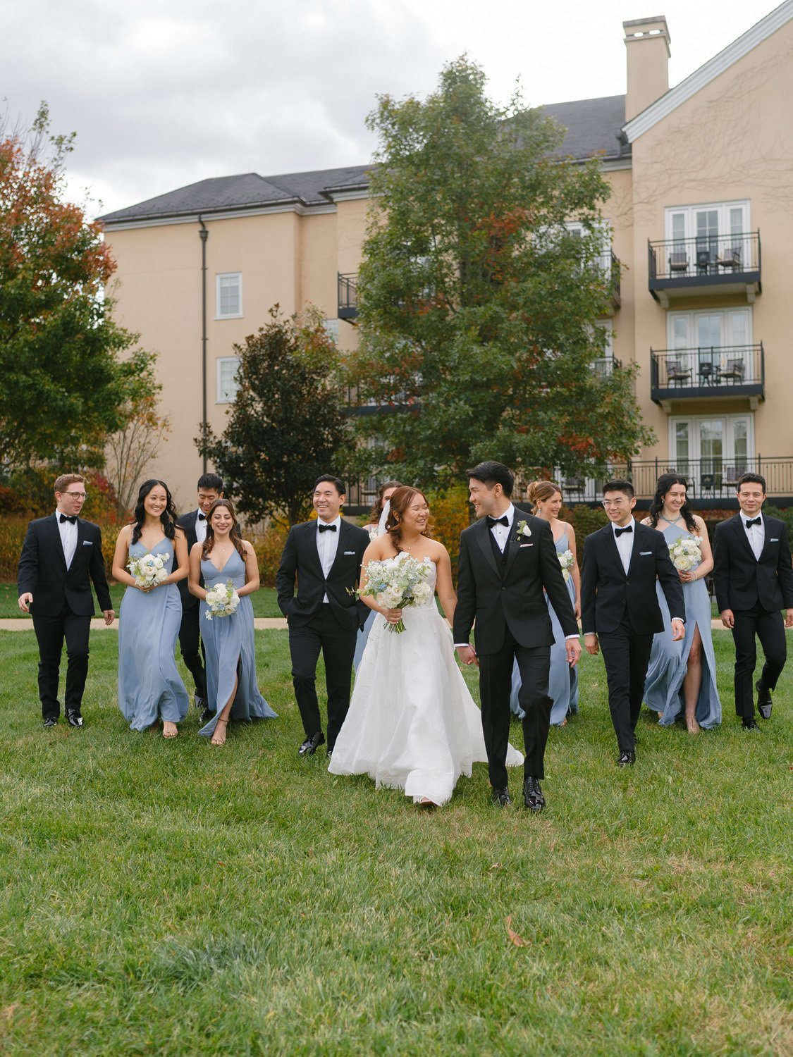 A wedding party walking on a grassy lawn with a building and trees in the background, featuring a bride and groom in the center, bride in a white wedding gown and groom in a black tuxedo, surrounded by bridesmaids in blue dresses and groomsmen in bla