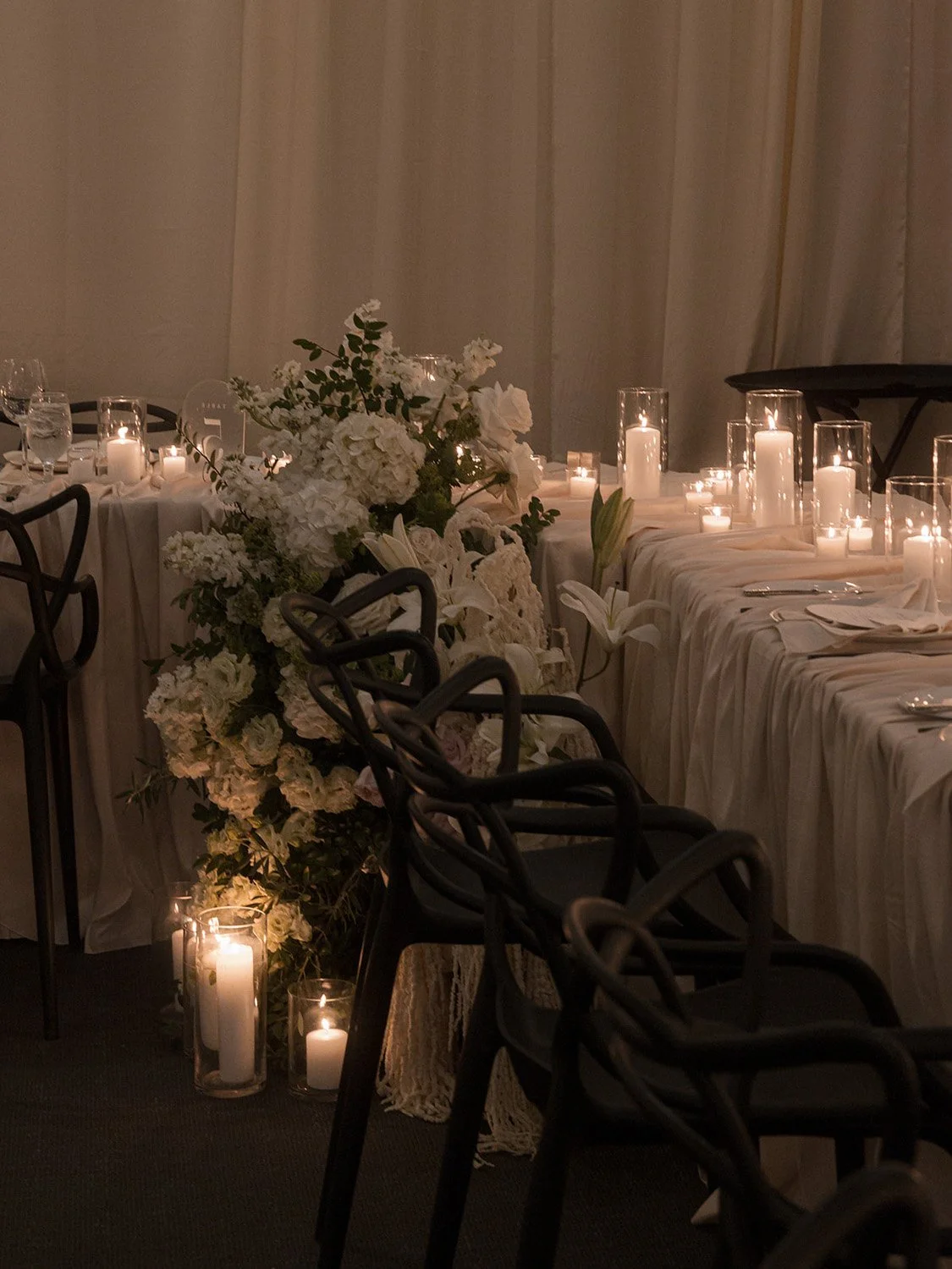 Elegant dining setup with black chairs, white tablecloths, floral arrangements, and numerous candles.