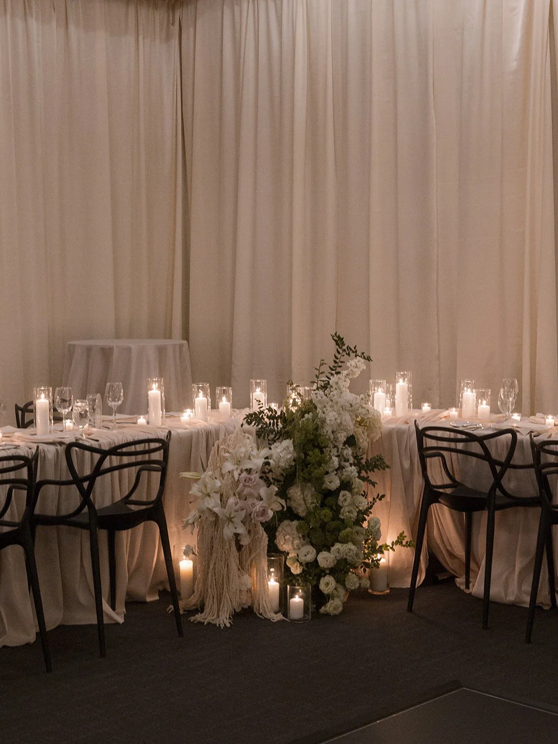 Elegant dining table decorated with white flowers, candles, and glassware, set against a cream-colored curtain backdrop in a formal event setting.