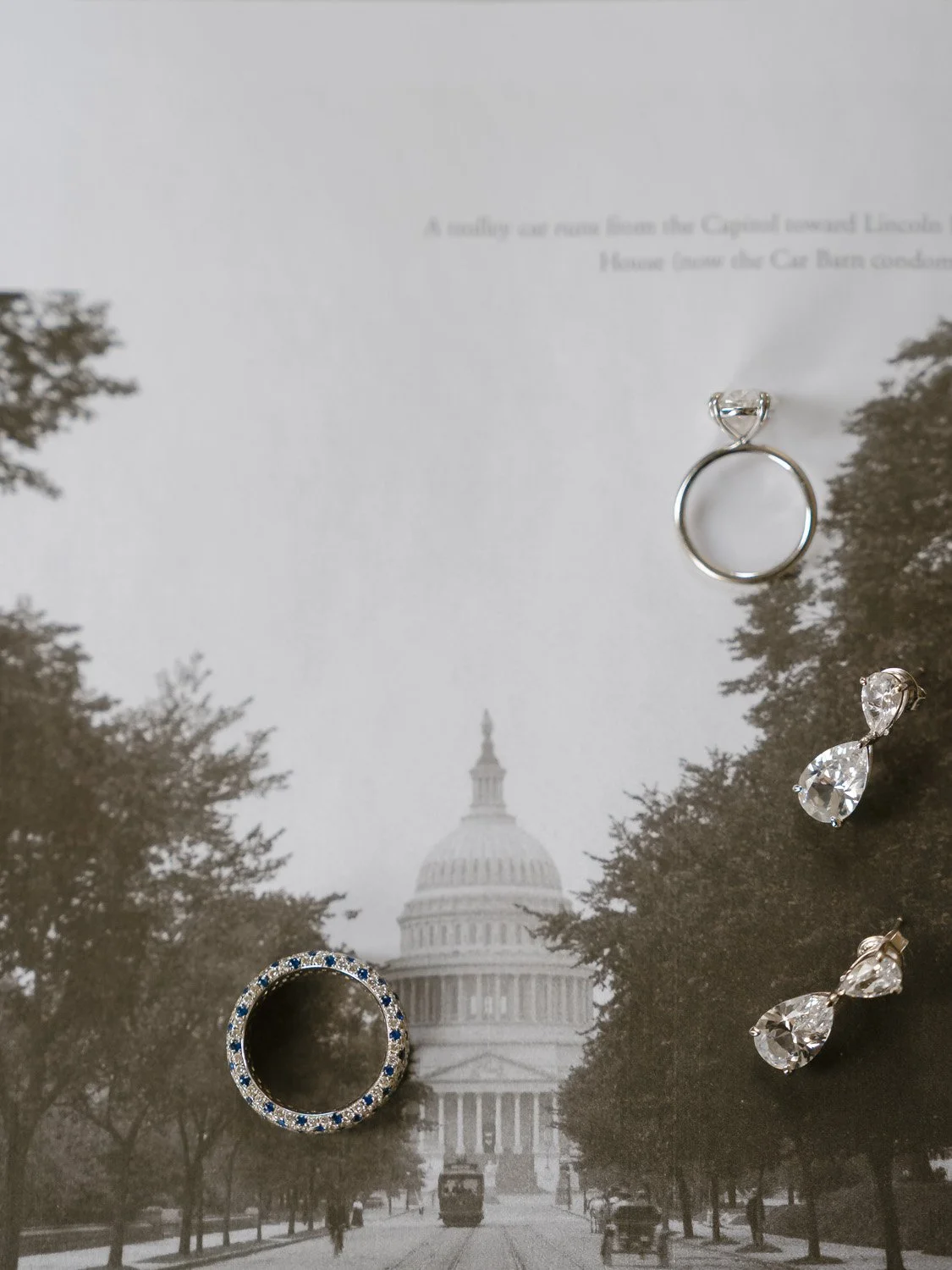 A black and white photograph of the U.S. Capitol building with trees lining the street, overlaid by several pieces of jewelry including an engagement ring, a pair of earrings, and a ring with blue stones.