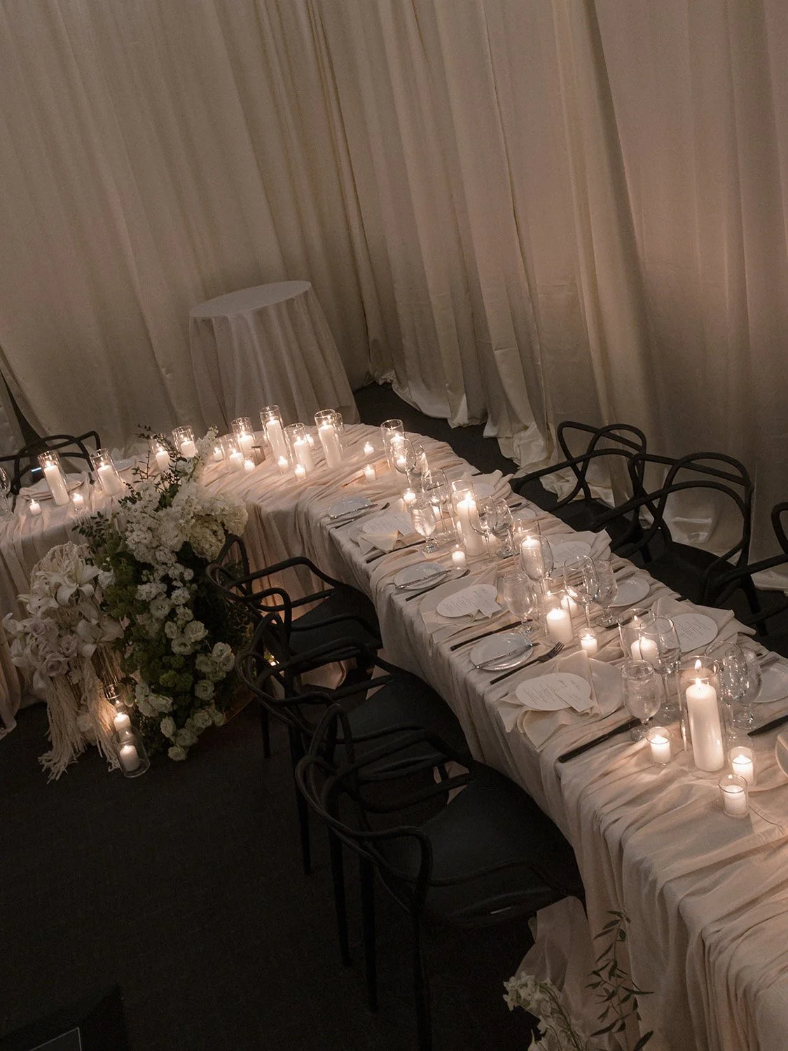 Elegant banquet table with candles, floral arrangement, and place settings, set for a formal event in a dimly lit room with draped white curtains.