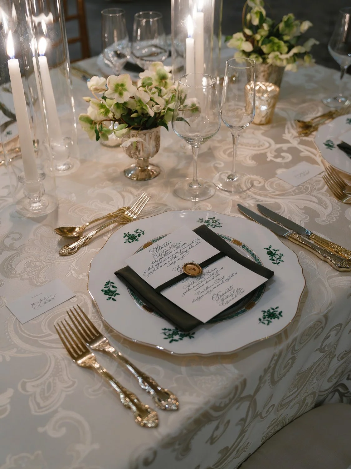 Elegant dining table setup with white-on-white patterned tablecloth, gold cutlery, white floral centerpieces, glassware, candles, place cards, and a menu on a plate.