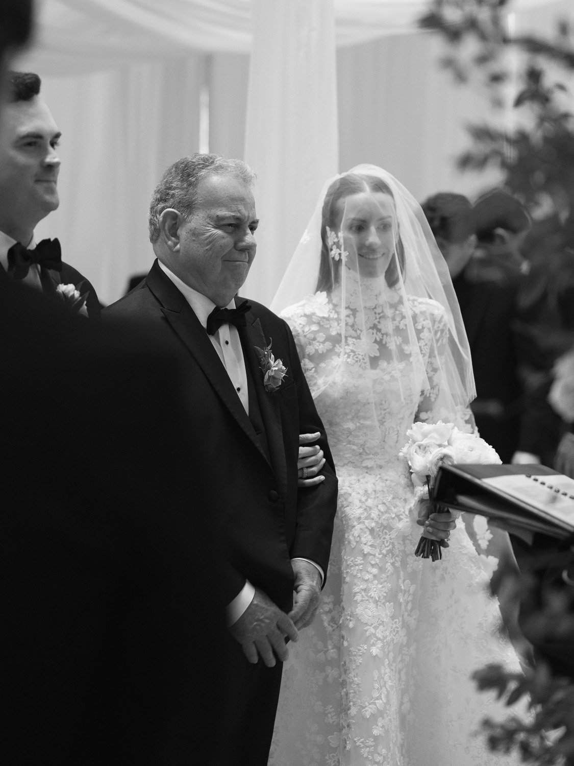 A black and white photo of a wedding ceremony, featuring a bride in a lace wedding gown and veil, standing next to a man in a tuxedo, with other people in the background.