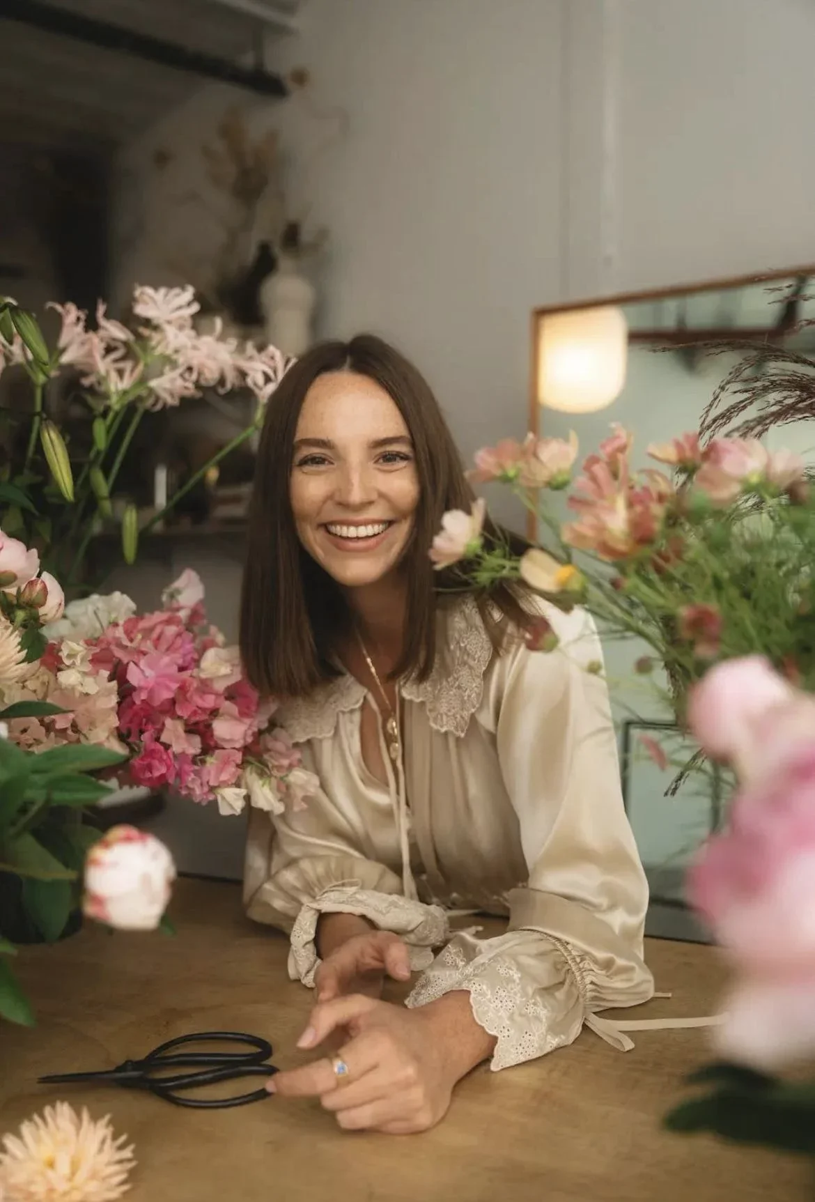A woman with shoulder-length brown hair smiling while surrounded by pink and white flowers on a table, with a cat sitting on a shelf in the background.