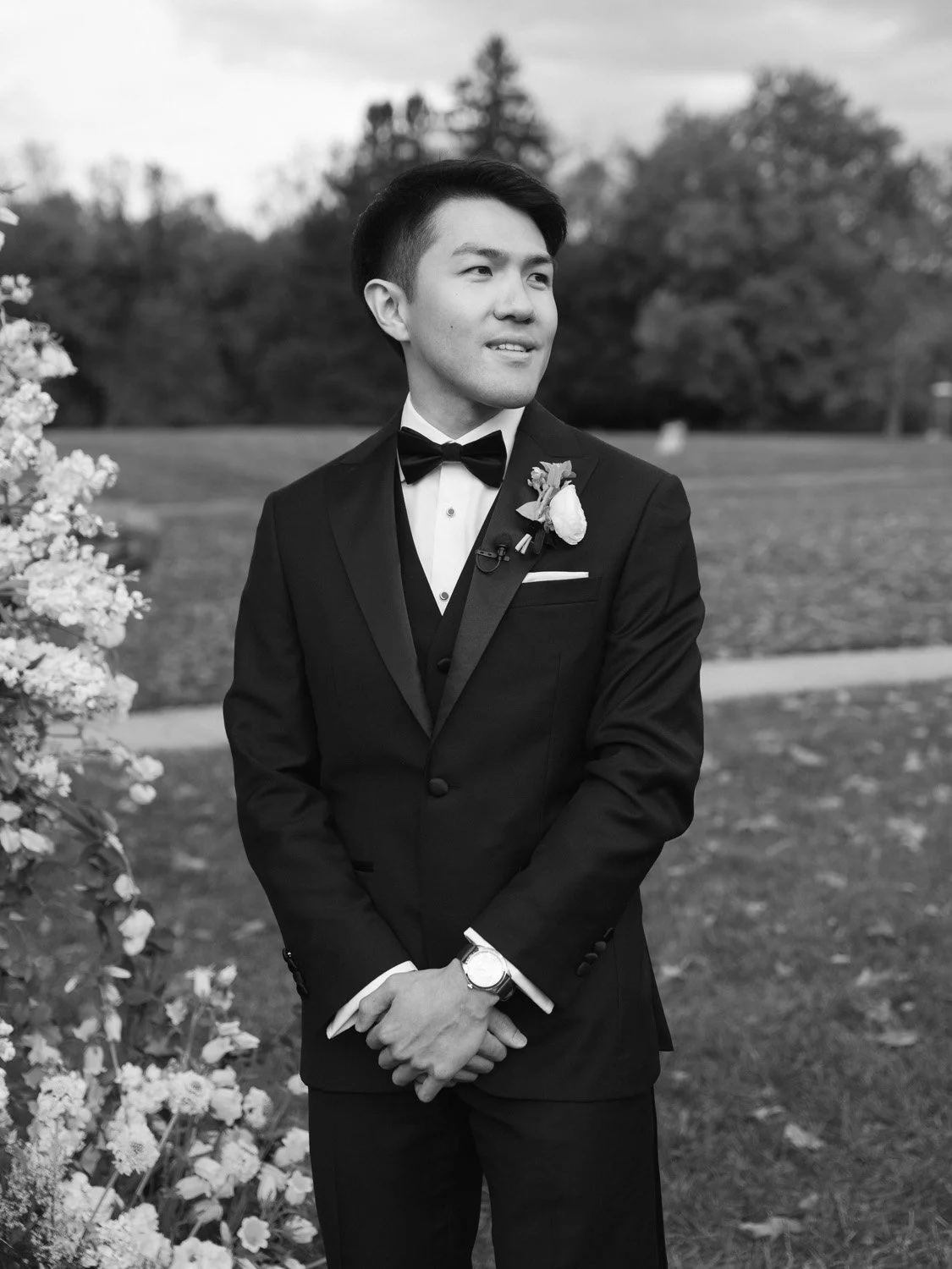Young man in tuxedo with bowtie, boutonniere, and wristwatch outdoors, smiling, with flower arrangement on one side and trees in the background.