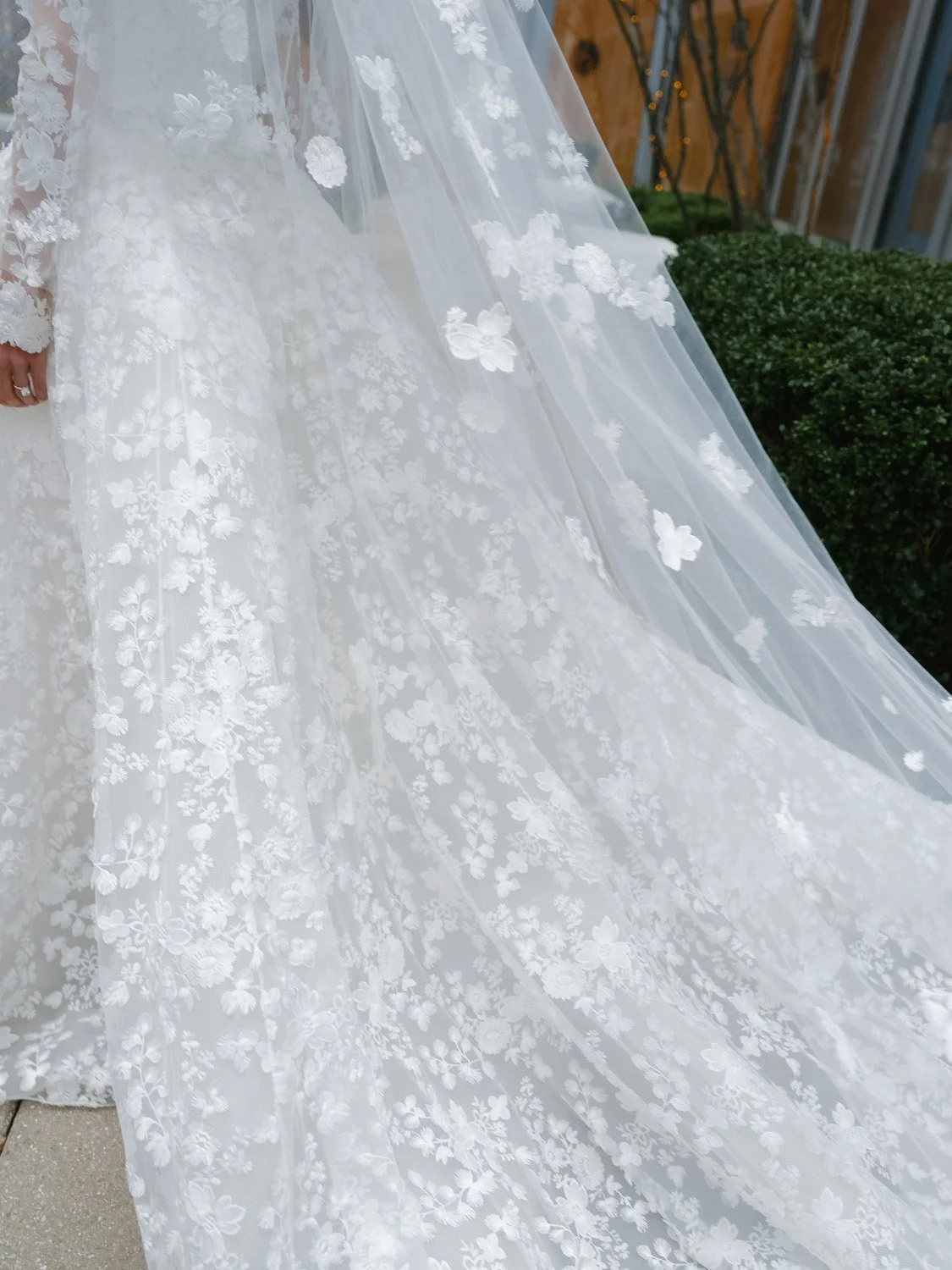 Close-up of a white wedding dress with floral lace embroidery and a flowing train, with a hand wearing a ring visible on the left side.