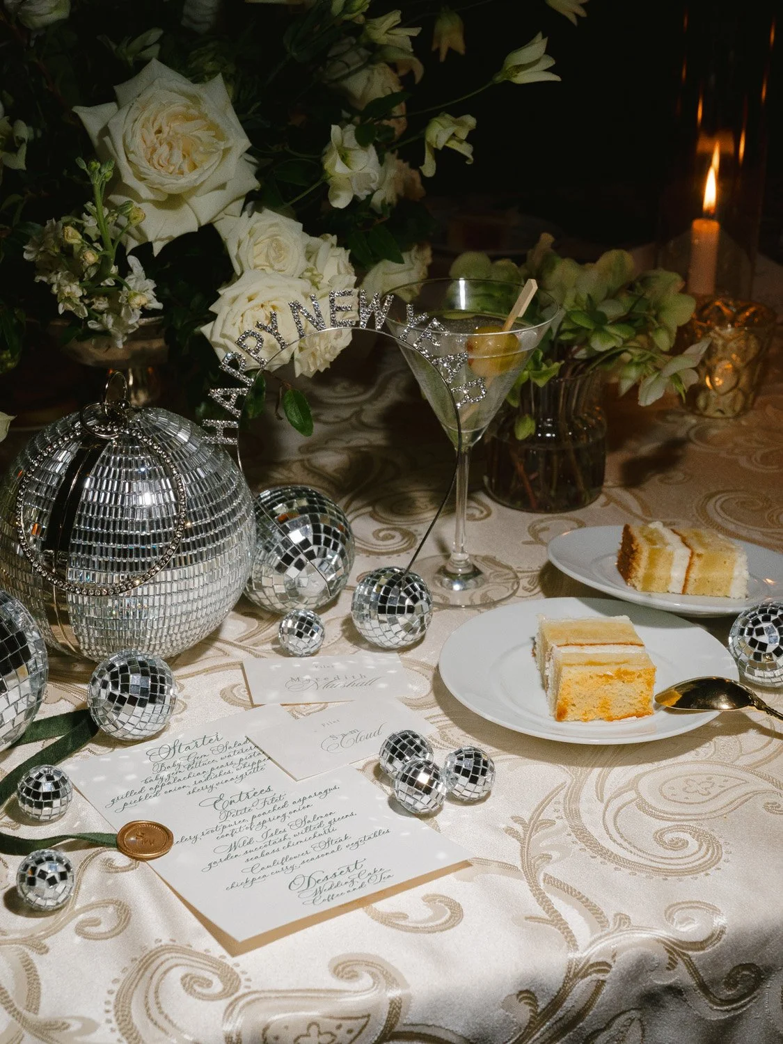 A decorated table with white roses, a disco ball, small disco ball ornaments, a cocktail glass, slices of cake on plates, a menu card, and a lit candle, indicating a celebration or party.