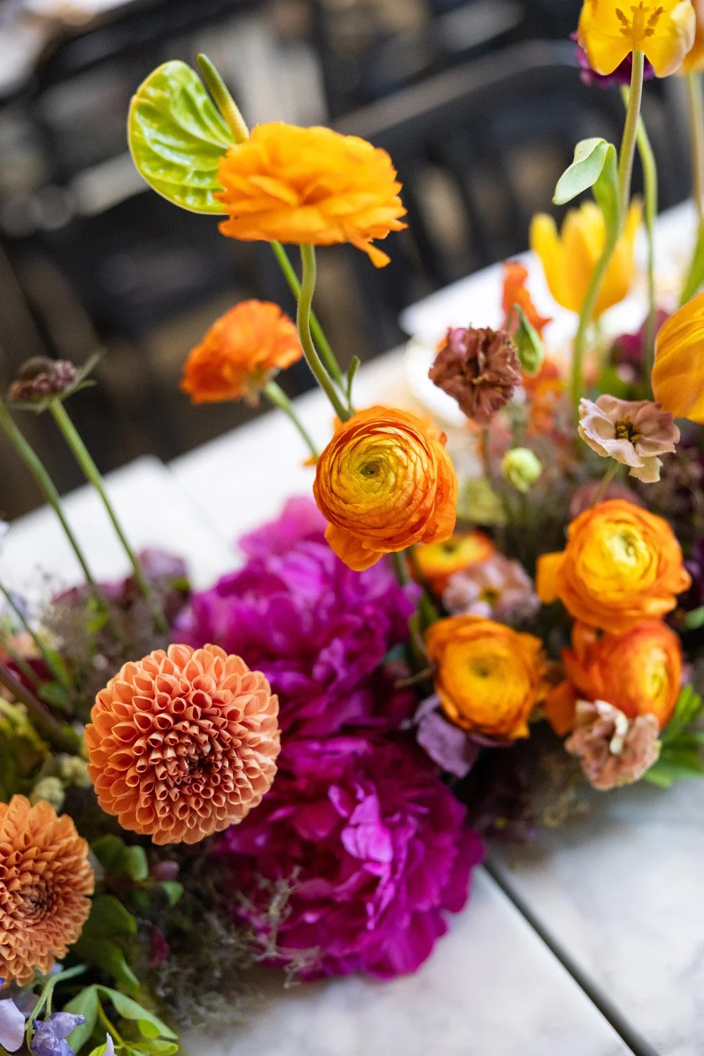 Colorful bouquet of various flowers, including orange ranunculus, pink dahlias, and yellow and purple flowers, arranged on a surface with a black fence in the background.