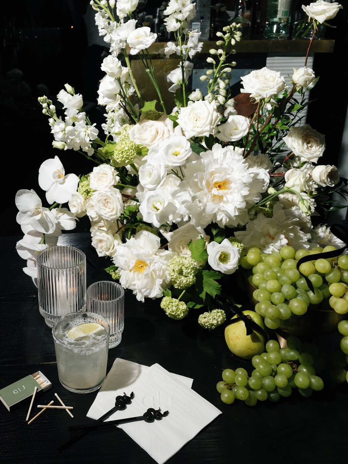 A floral arrangement with white roses, peonies, and orchids, along with green grapes and an apple, placed on a dark table with candles and drink glasses.