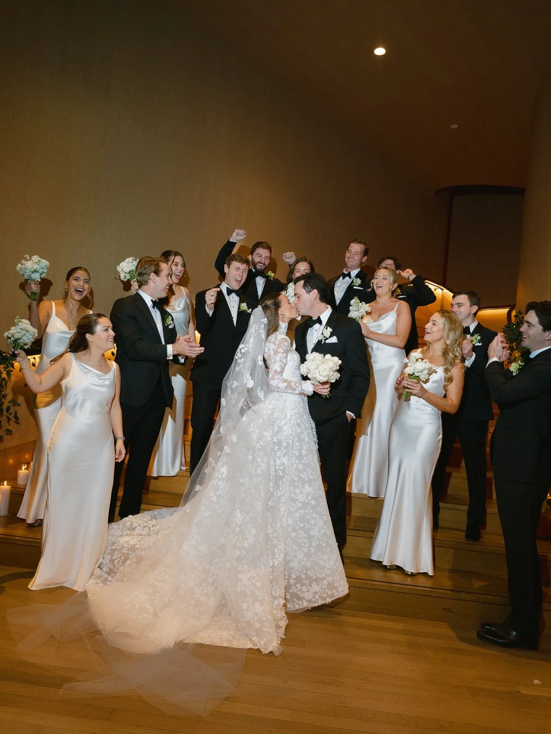 A wedding party celebrating, with a bride and groom kissing surrounded by bridesmaids and groomsmen in formal attire.