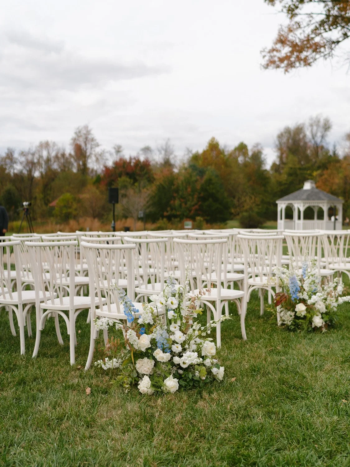 Outdoor wedding setup with white chairs and floral arrangements, overcast sky, trees in fall colors, and a white gazebo in the background.