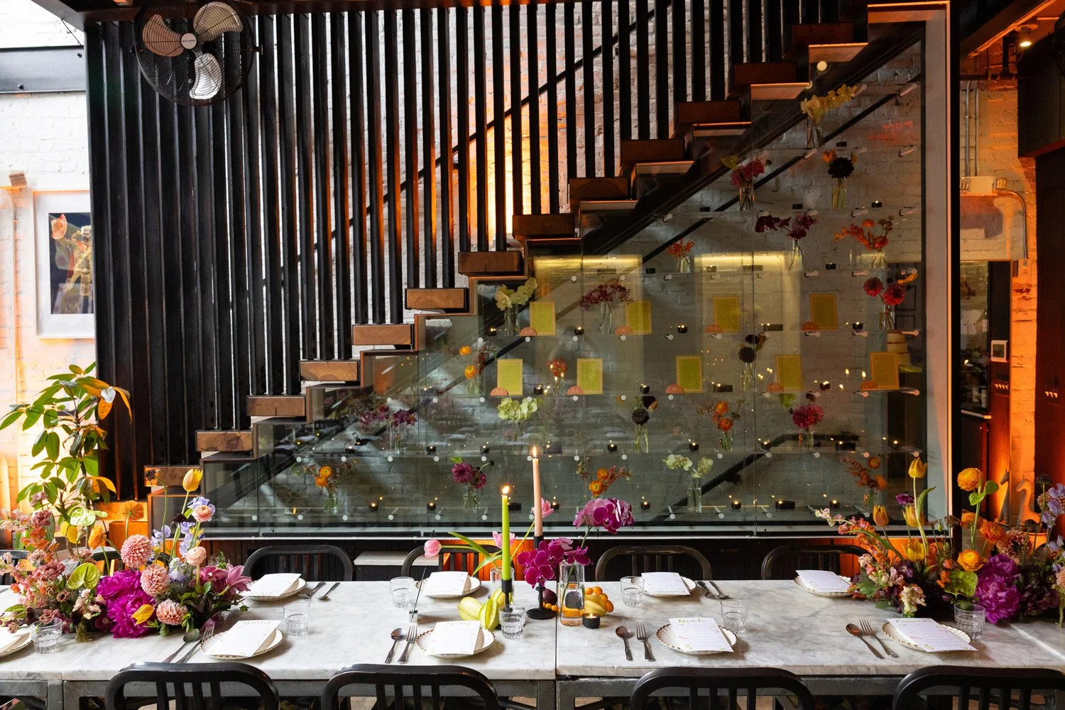Indoor dining area with a large marble table, floral centerpieces, candles, and place settings, with a glass wall overlooking a staircase decorated with flowers.