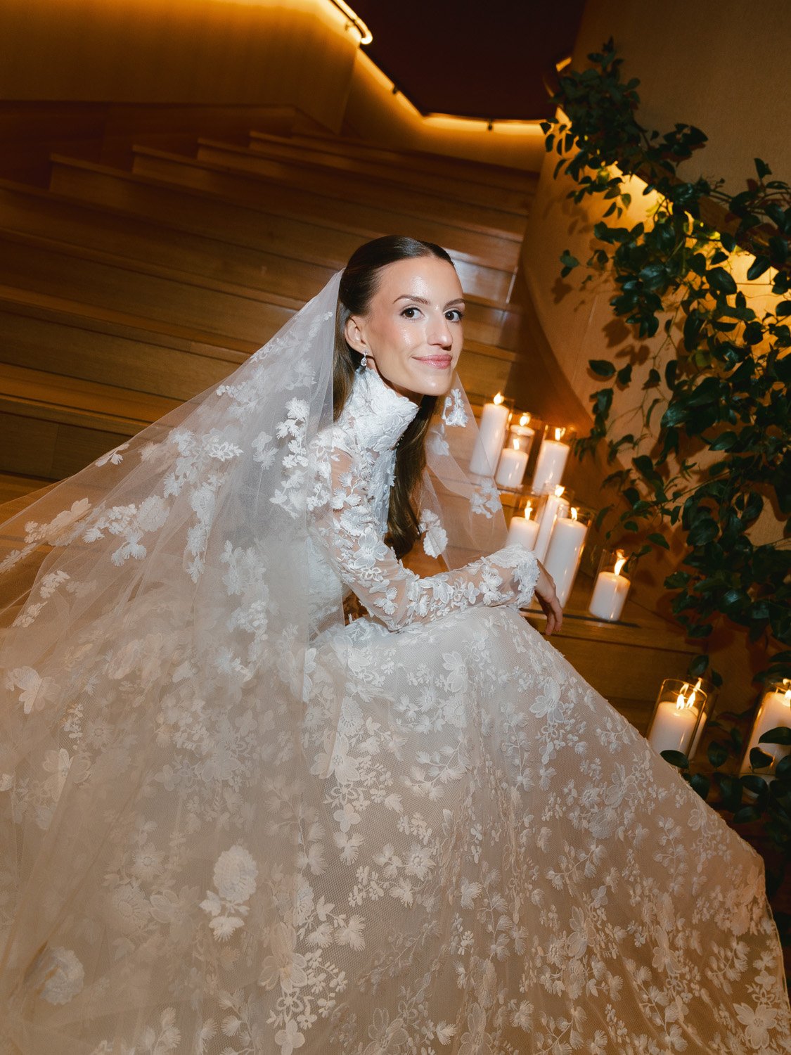 A bride sitting on wooden stairs, wearing a lace white wedding gown and veil, with lit candles in the background and greenery nearby.