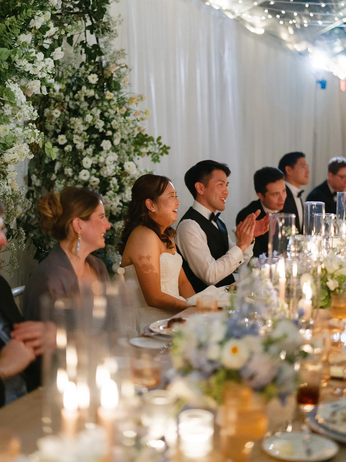 People sitting at a wedding reception table, smiling and clapping, with floral decorations and candles.