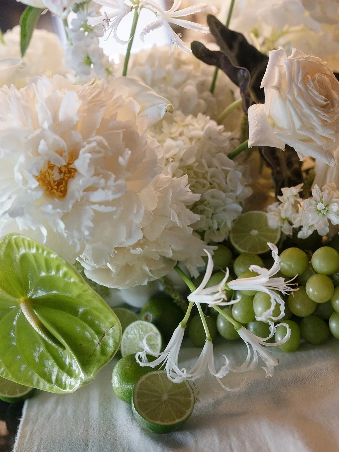 Close-up of a floral arrangement featuring white peonies, white lilies, white daisies, green anthuriums, and limes, some sliced open showing the interior.