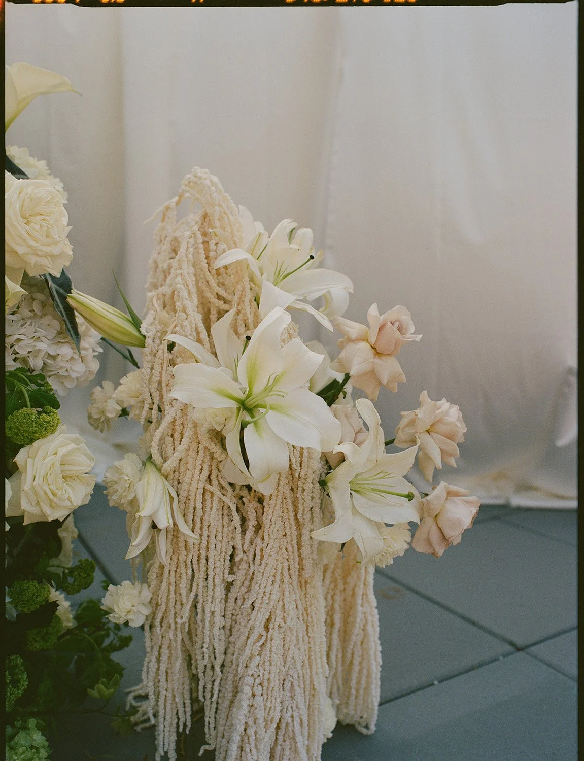A floral arrangement featuring white lilies, roses, and other flowers, with a backdrop of white drapery.