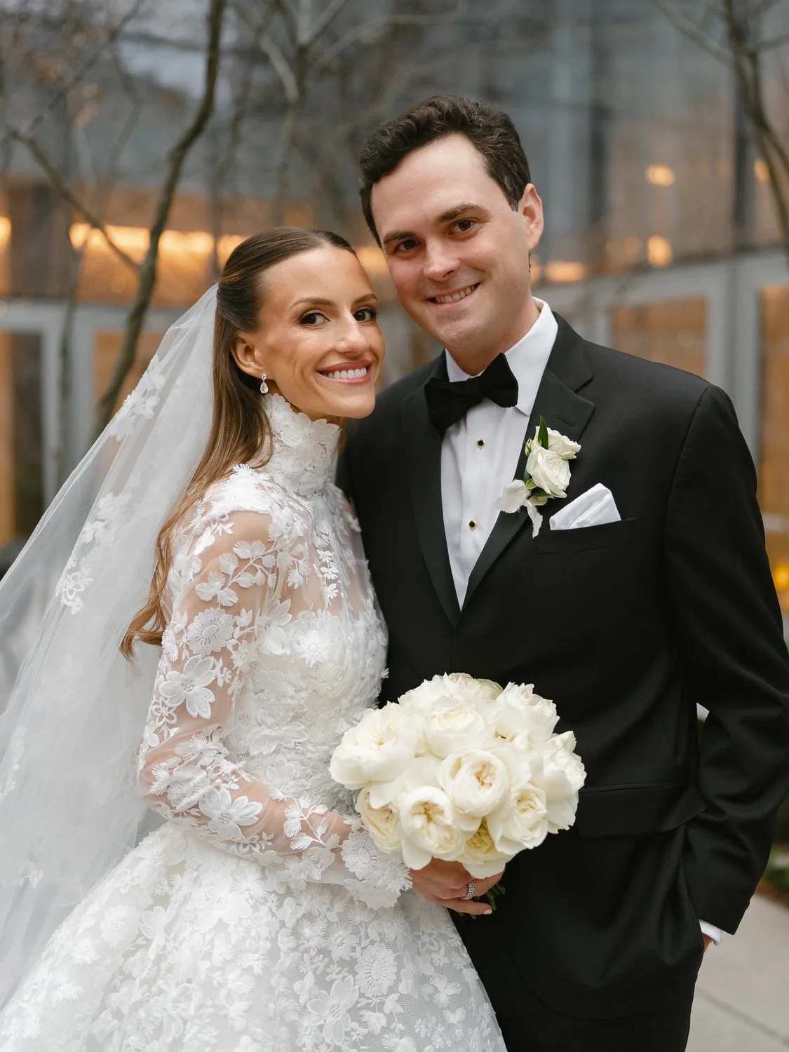 A bride and groom smiling and posing together on their wedding day, with the bride holding a bouquet of white flowers and wearing a lace wedding dress, and the groom in a black tuxedo with a white shirt and black bow tie.
