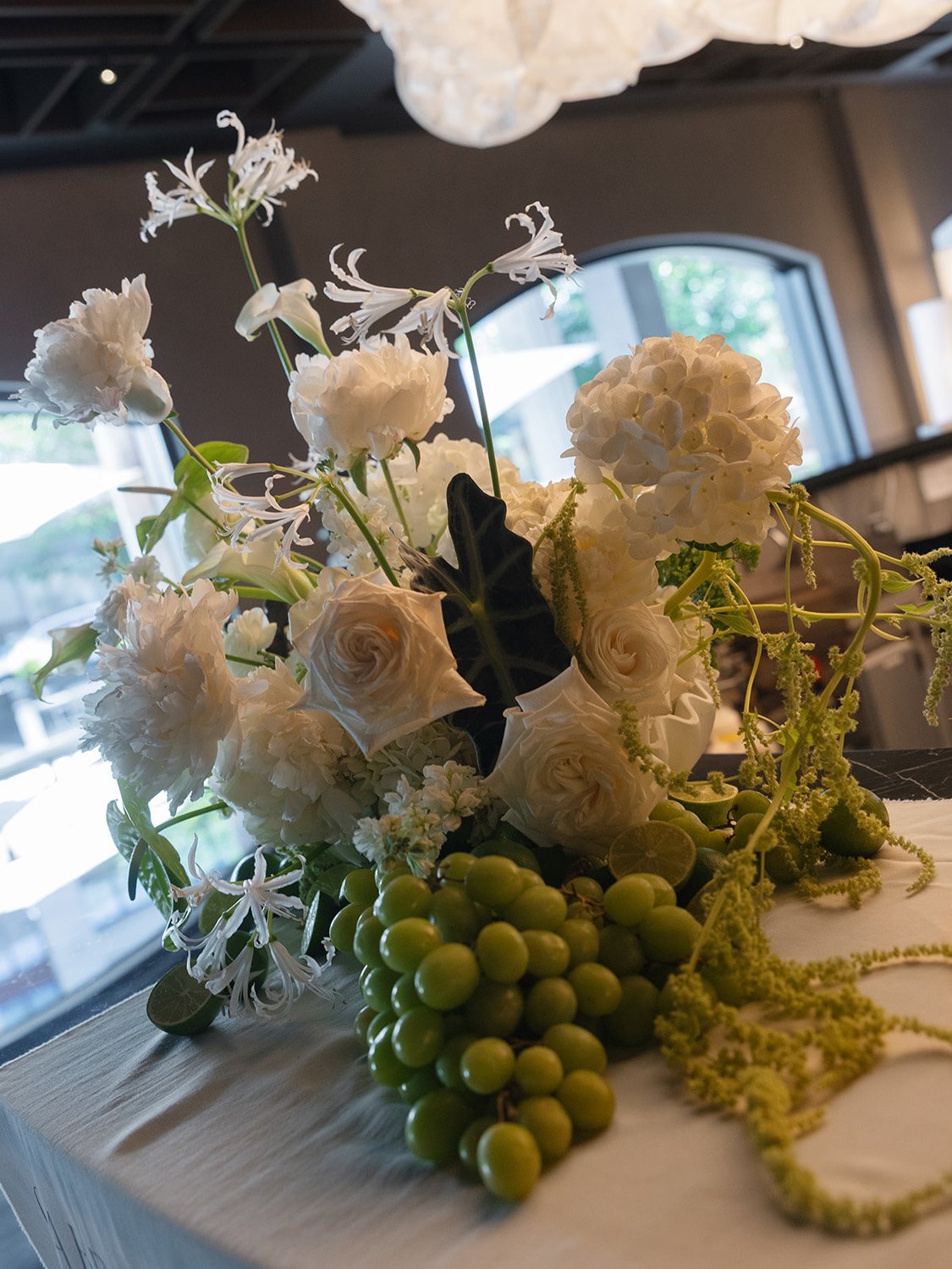 White floral arrangement with roses, hydrangeas, and greenery on a wooden surface near a window.