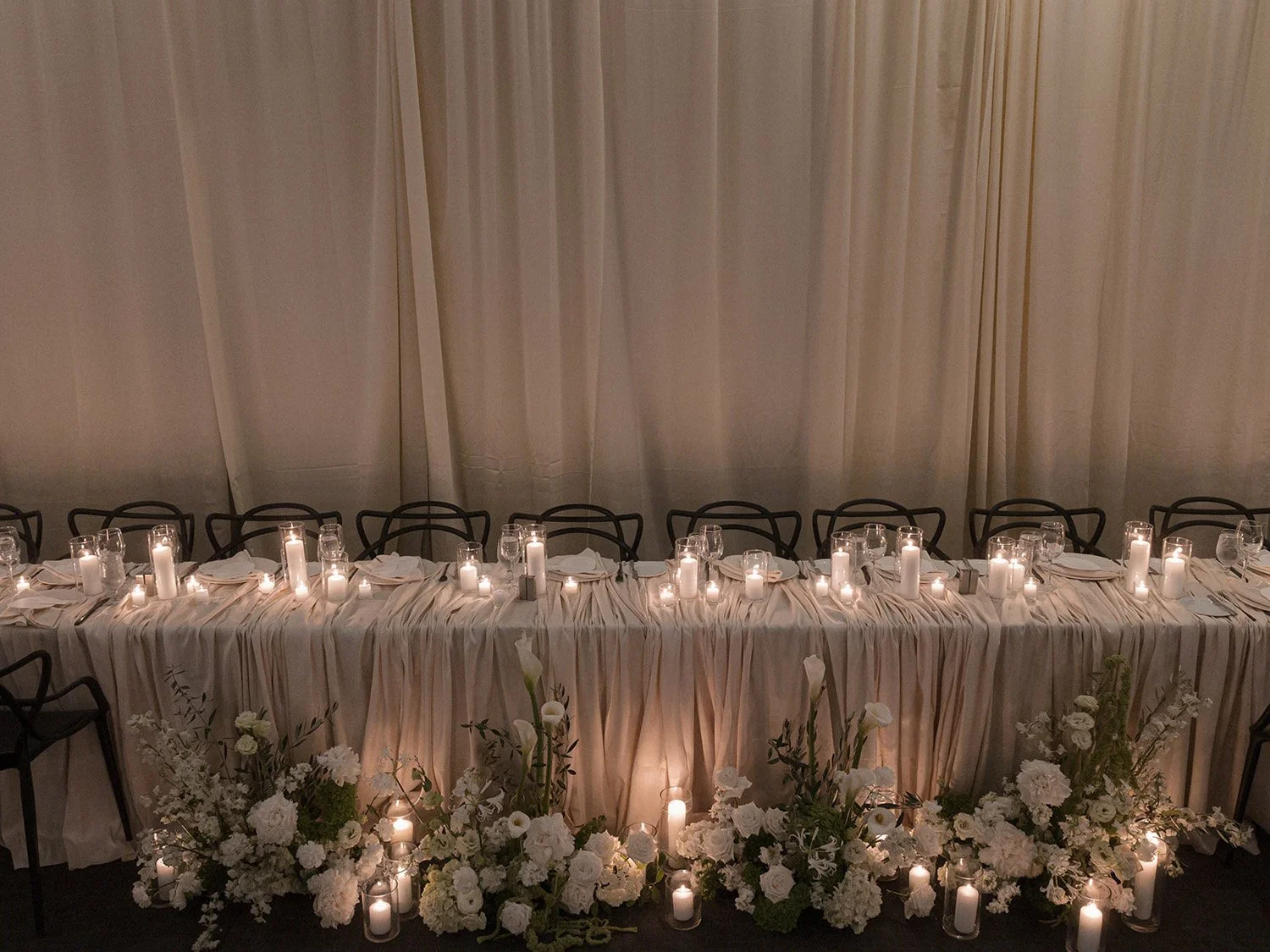 Elegant banquet table with white tablecloth, candles, glassware, and floral arrangements under a white curtain backdrop.