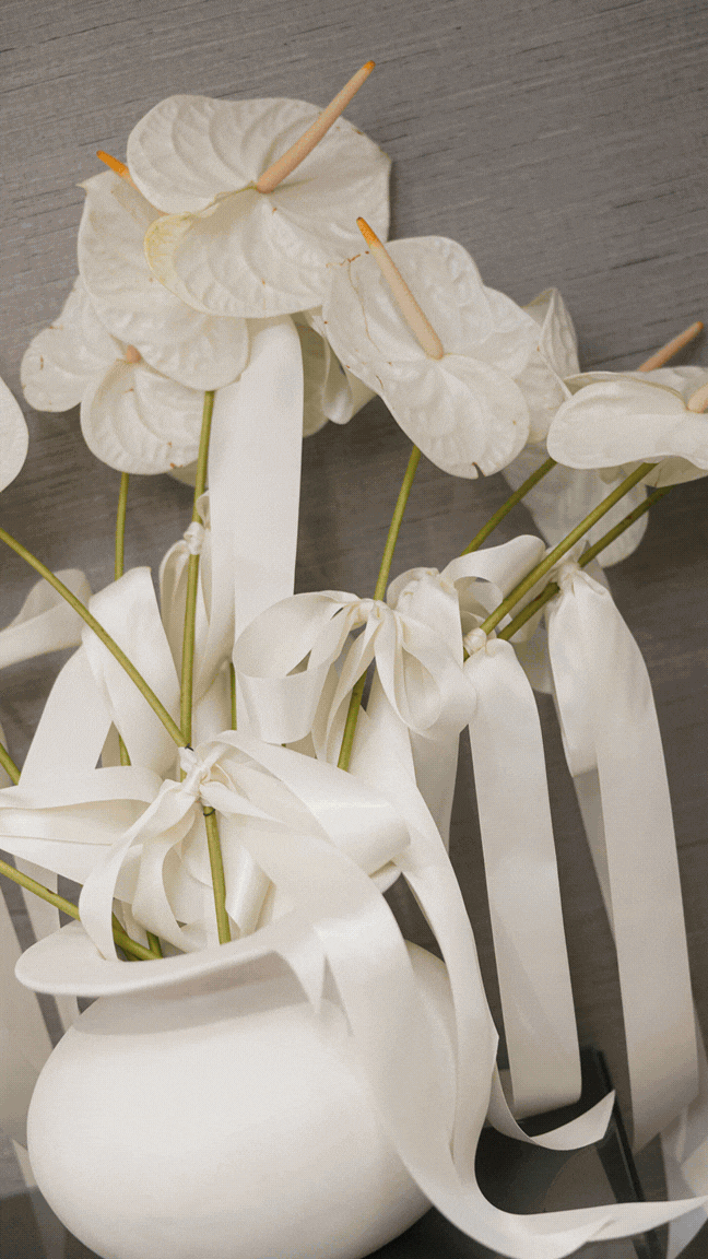 White anthurium flowers with ribbons in a white vase on a gray wooden surface.