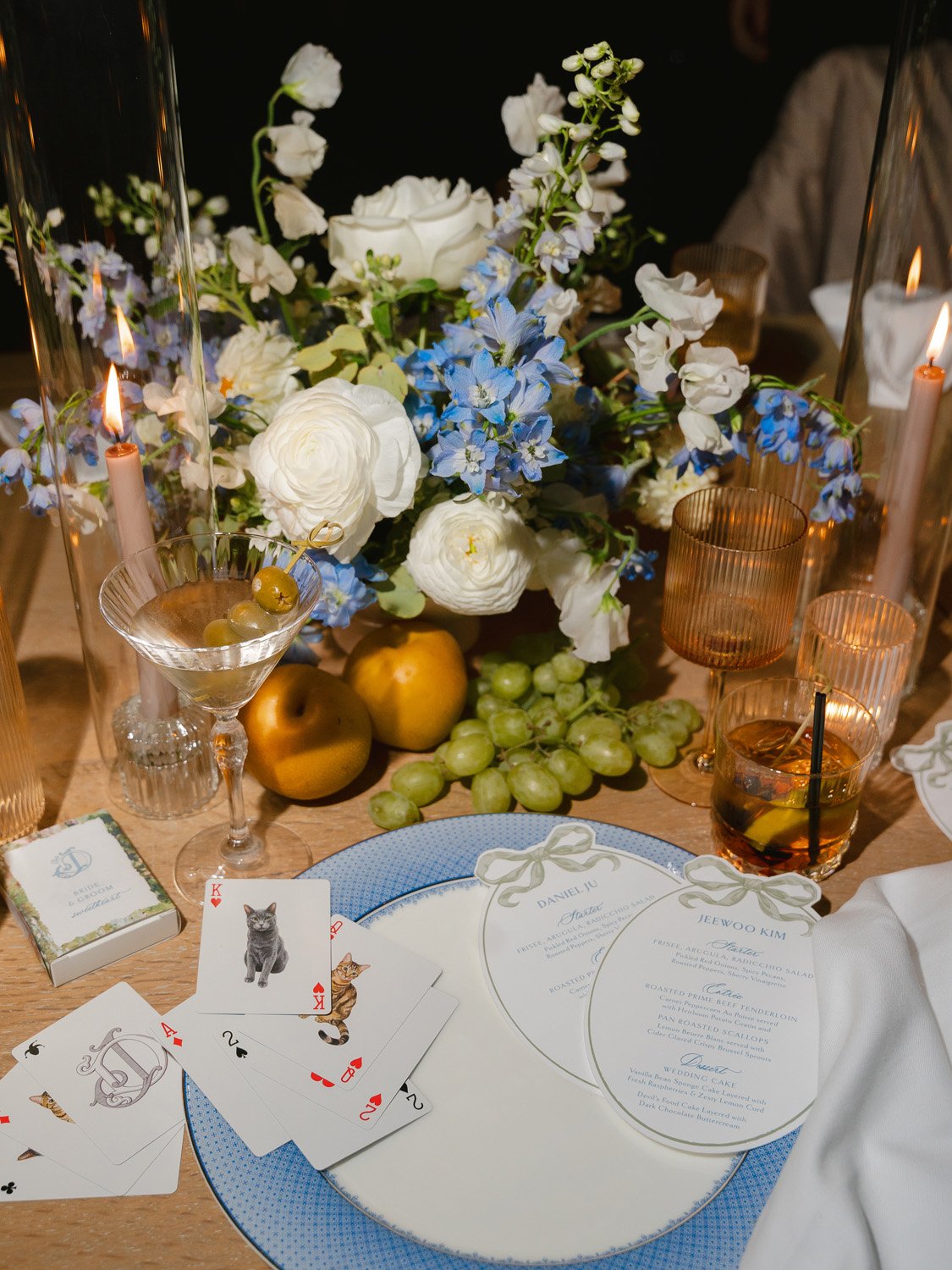 A table setting featuring a floral centerpiece with white and blue flowers, a card deck with playing cards, and drinks in glasses surrounded by candles.