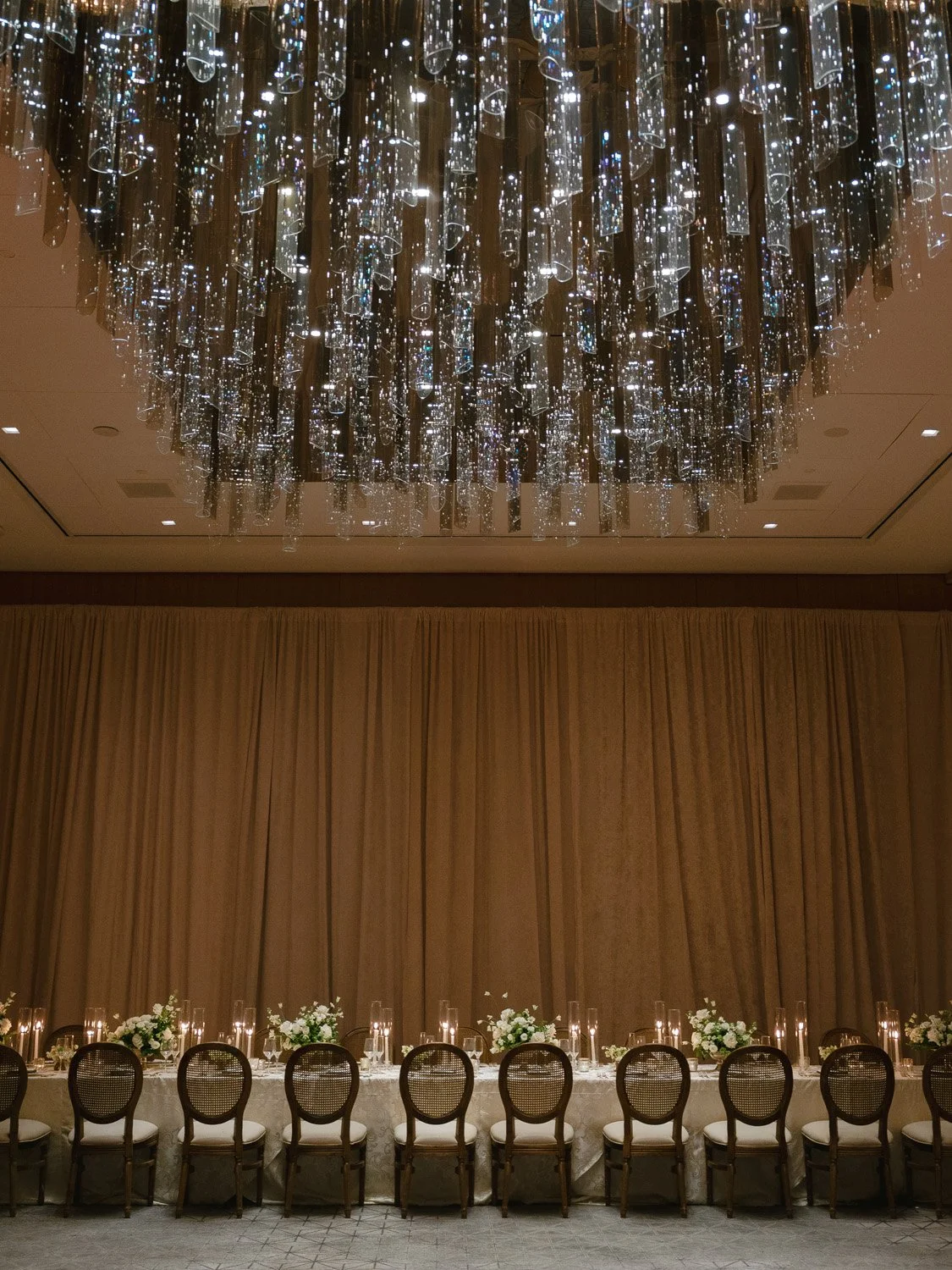 Elegant banquet table set with floral centerpieces and candles, ornate chairs, gold curtains, and a large modern chandelier with hanging glass or acrylic tubes.