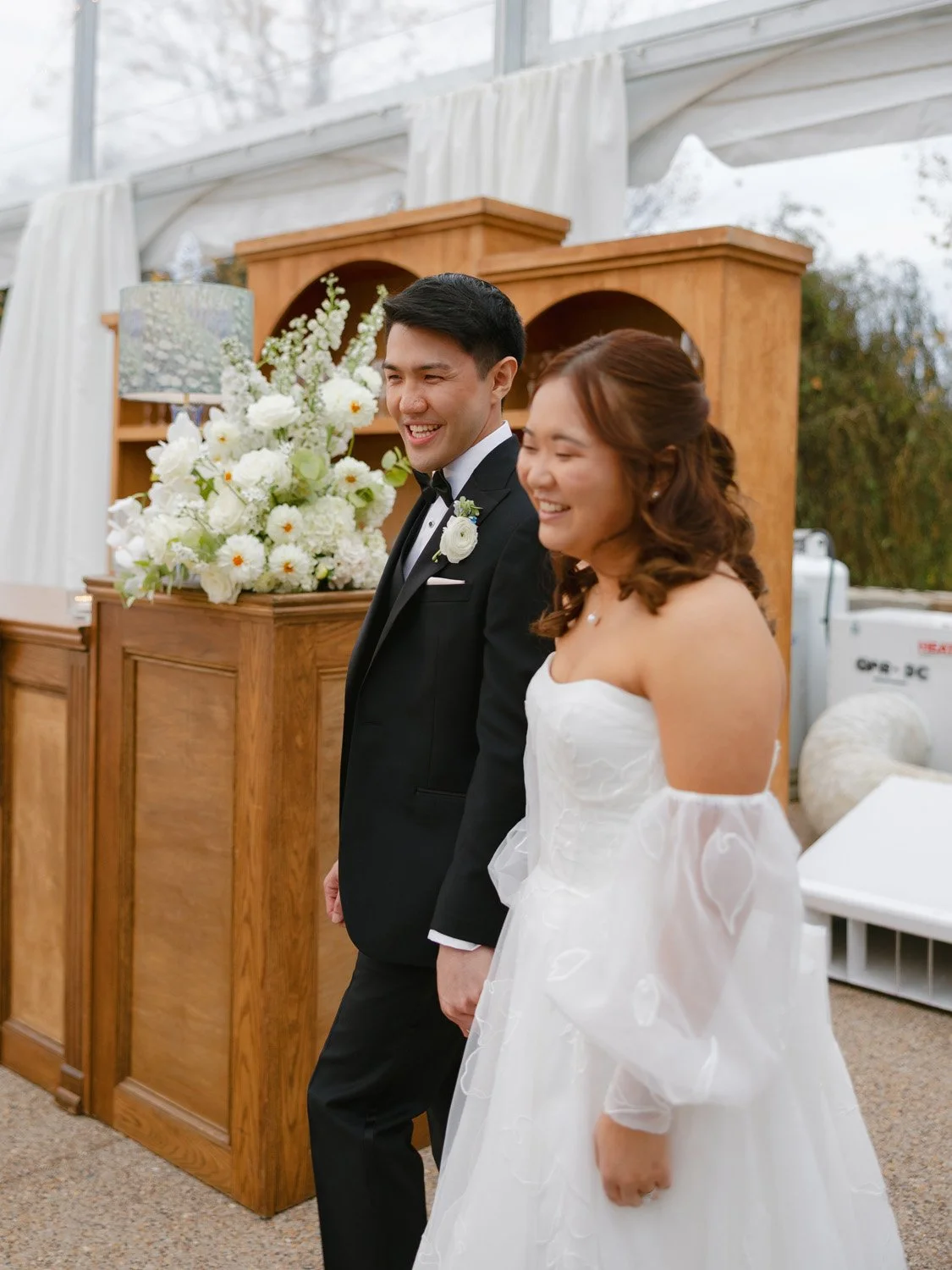 A bride and groom standing side by side, smiling during their wedding ceremony, in an indoor venue with floral arrangements and wooden furniture.