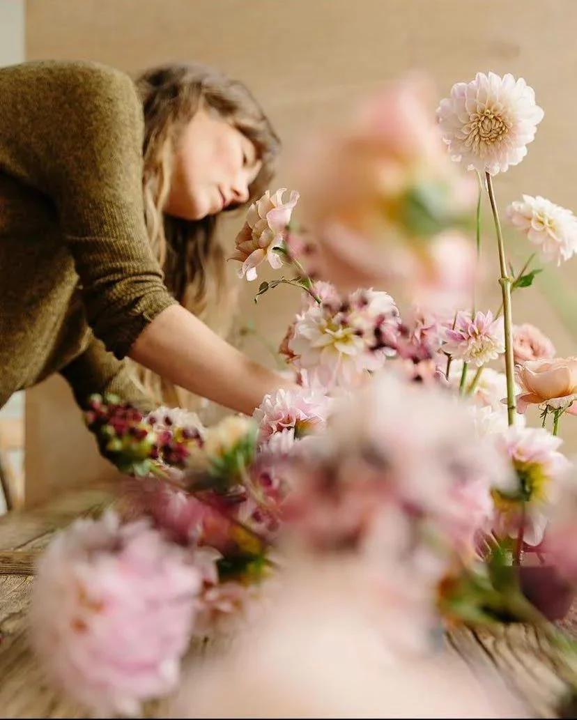 A woman arranging pink and white flowers on a table, with some flowers overlapping in the foreground.