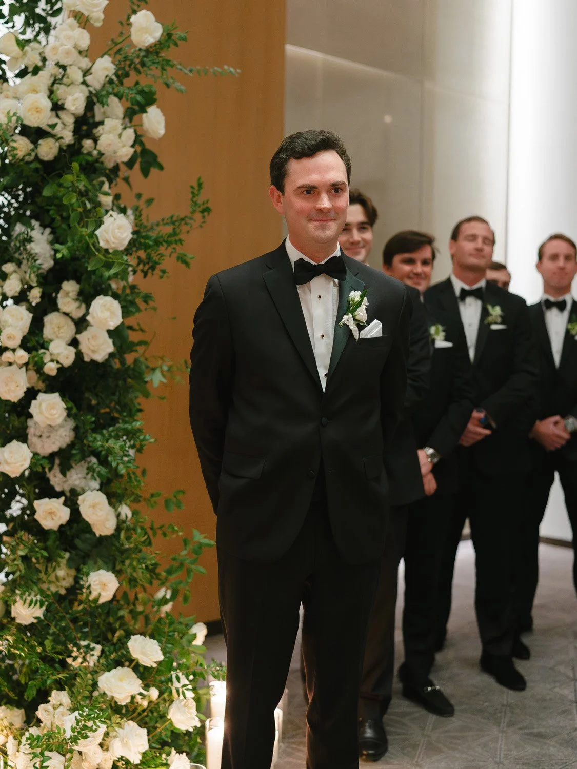 A man in a black tuxedo with a bow tie, standing with hands behind his back, during a wedding ceremony. Several men in similar attire are standing behind him, and a floral arrangement with white flowers is visible on the left side of the image.